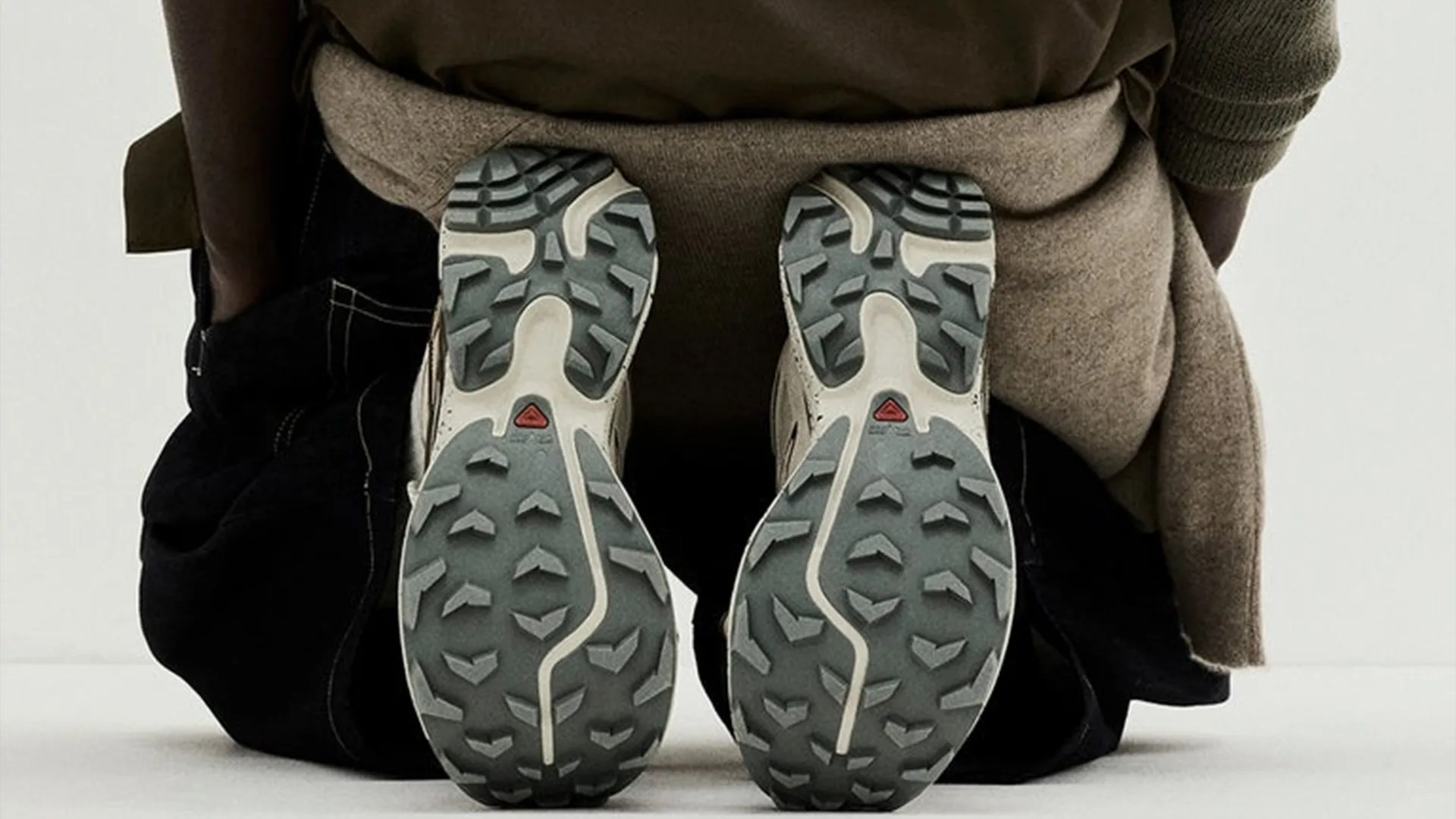 Pair of beige and gray hiking shoes with rugged soles worn by a person kneeling on a white surface.