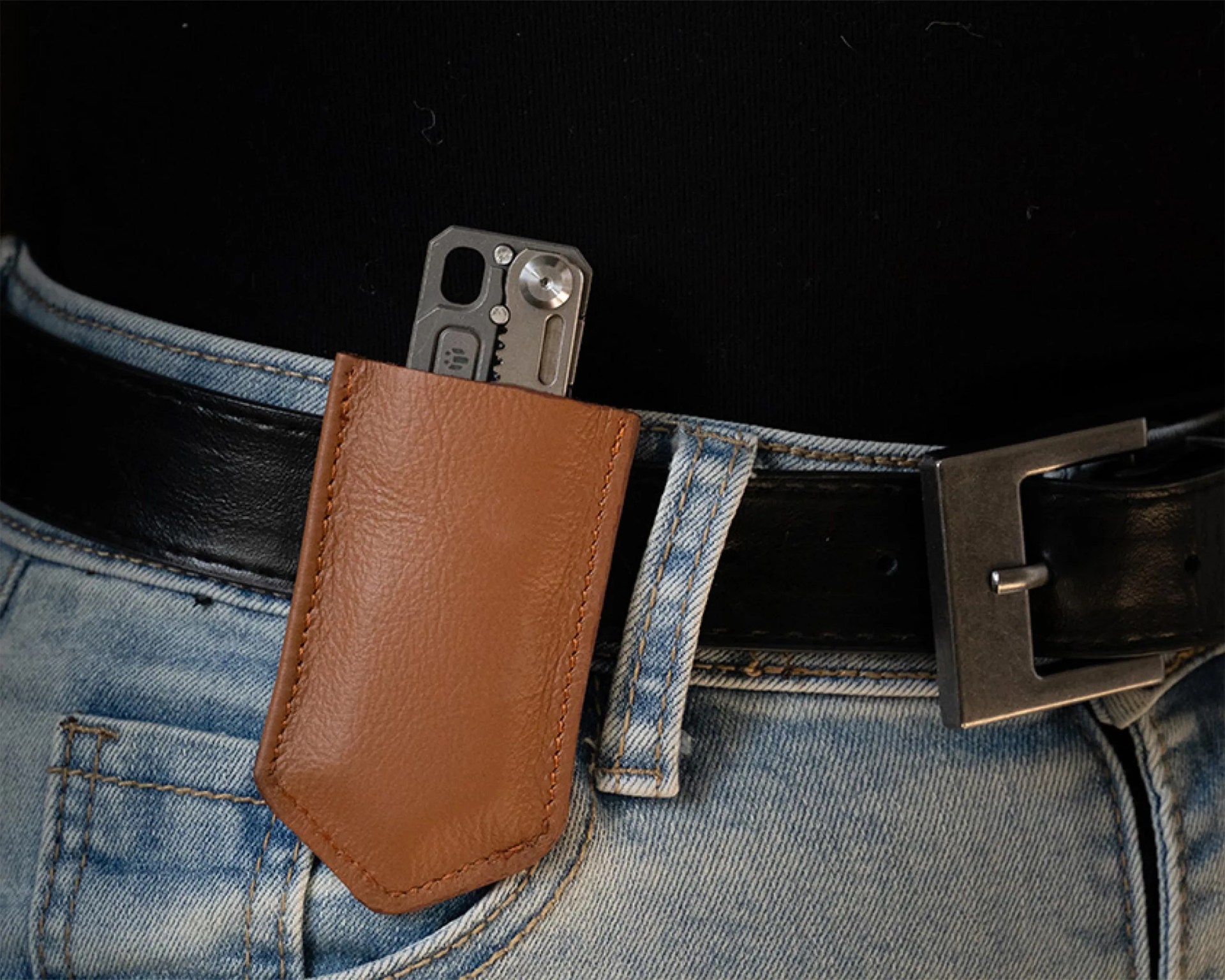 Brown leather pocket sheath holding a metal multi-tool clipped to a black belt on blue jeans.