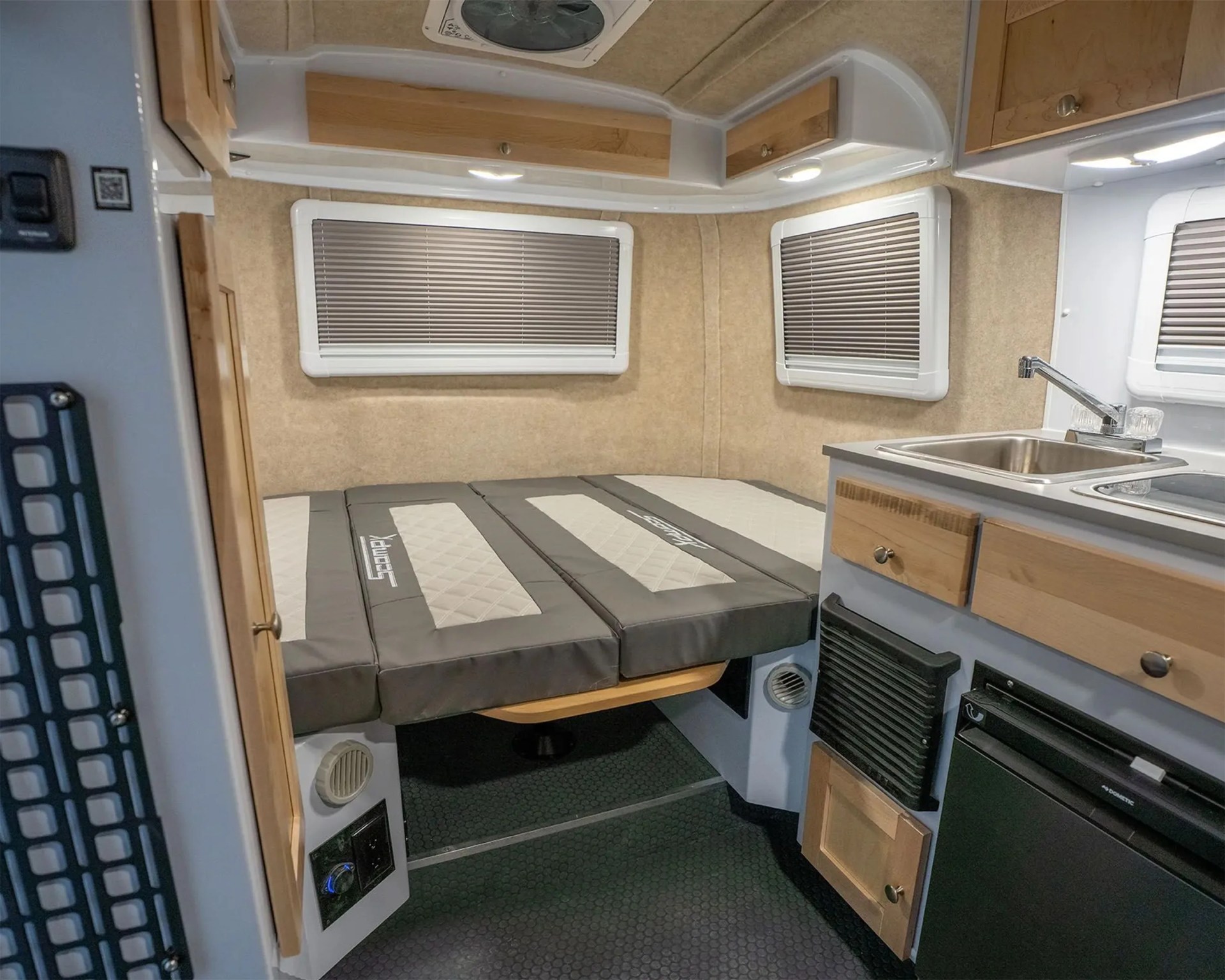 Compact camper interior with a foldable gray and beige bed, wooden cabinets, and a small kitchen sink and fridge.