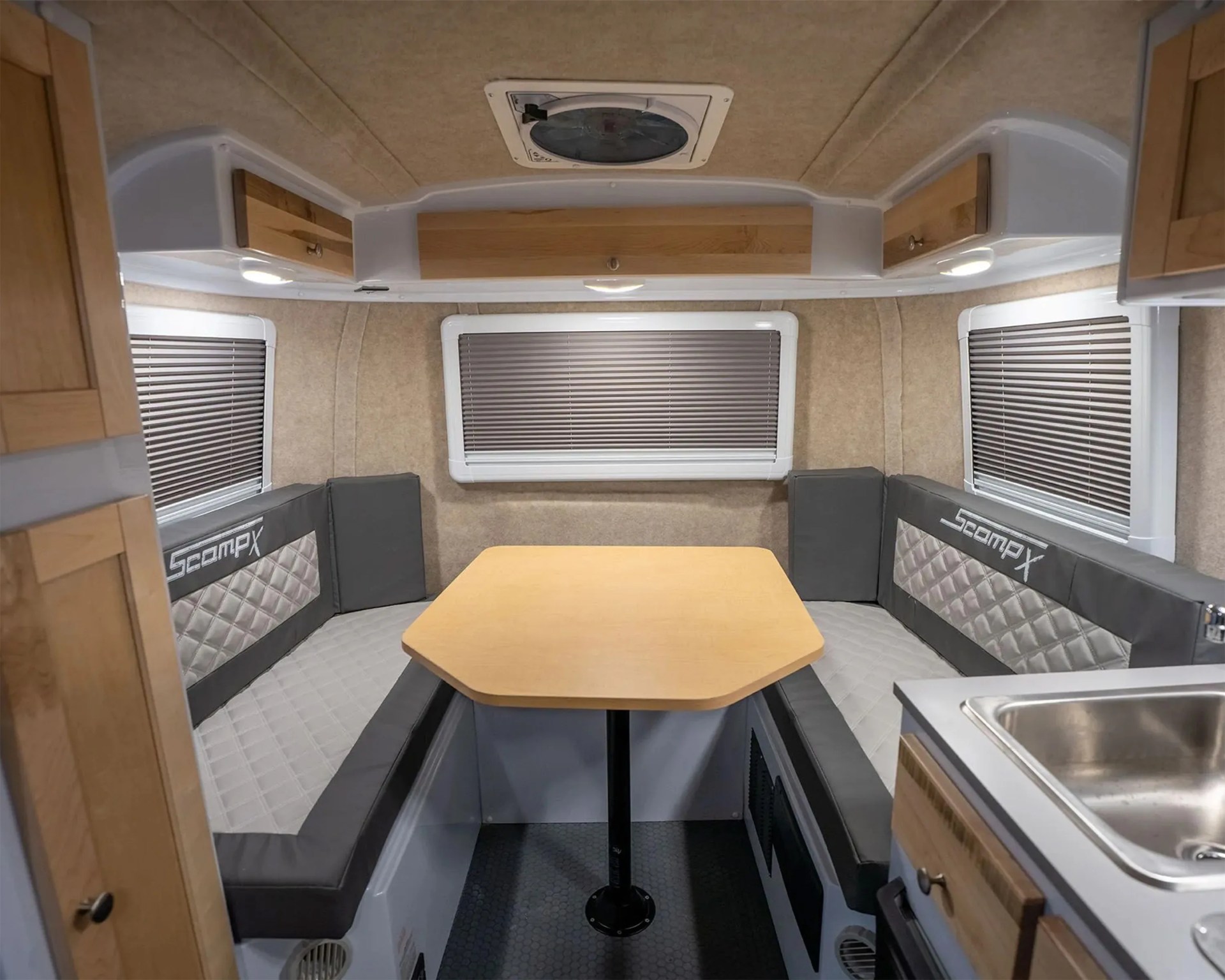Compact camper dinette area with a light wood table, gray cushioned bench seats, and windows with blinds.