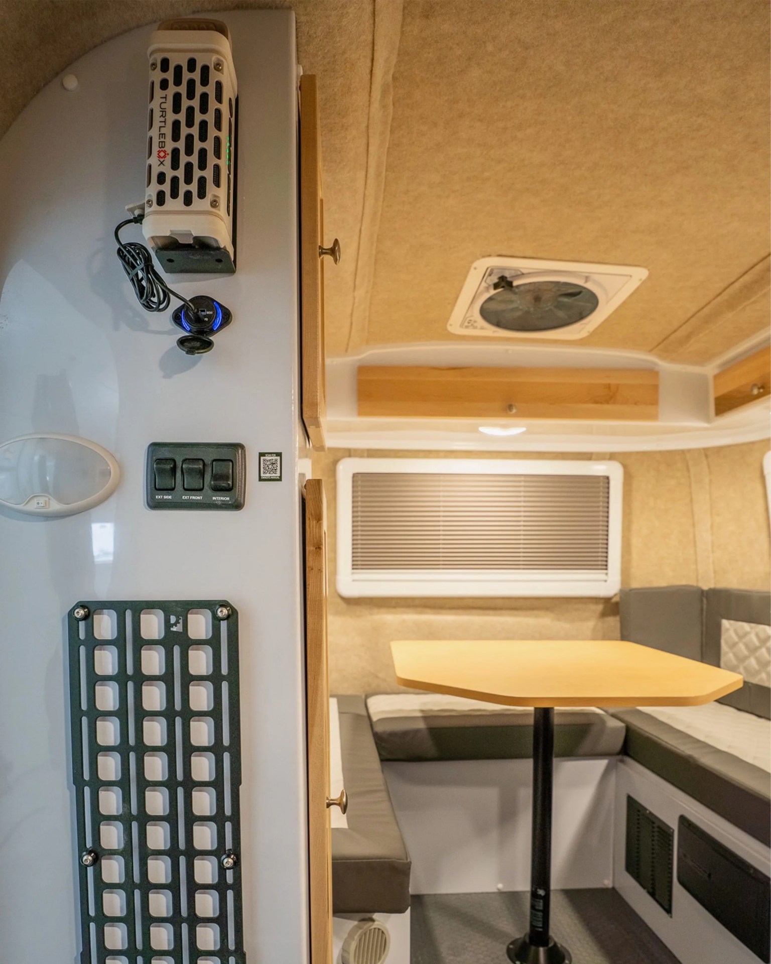 Compact camper interior with a small wooden table, cushioned bench seating, control switches, and a mounted Turtlebox device.