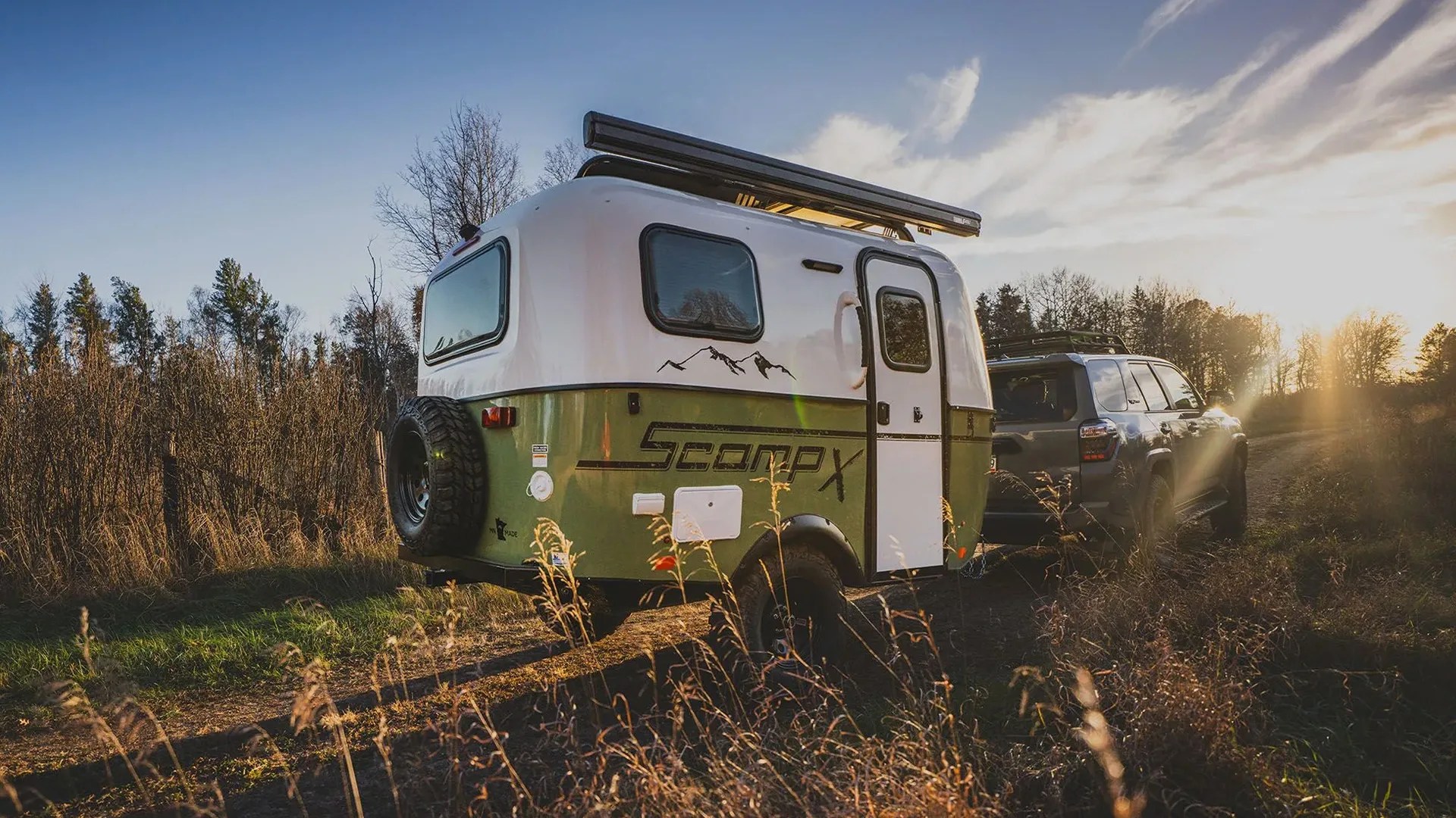Green and white Scamp X camper trailer hitched to a gray SUV on a dirt path at sunset.