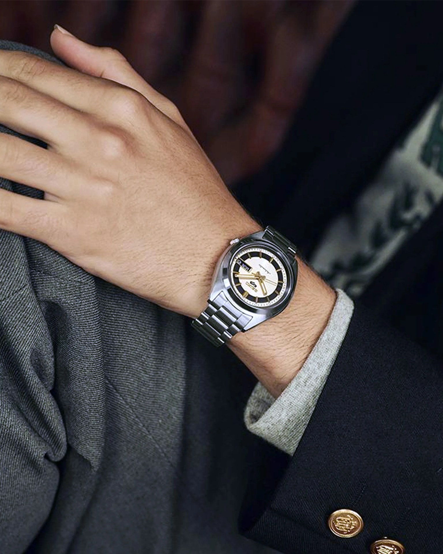 Silver metal wristwatch with a white and black dial worn on a wrist over a gray and black jacket.