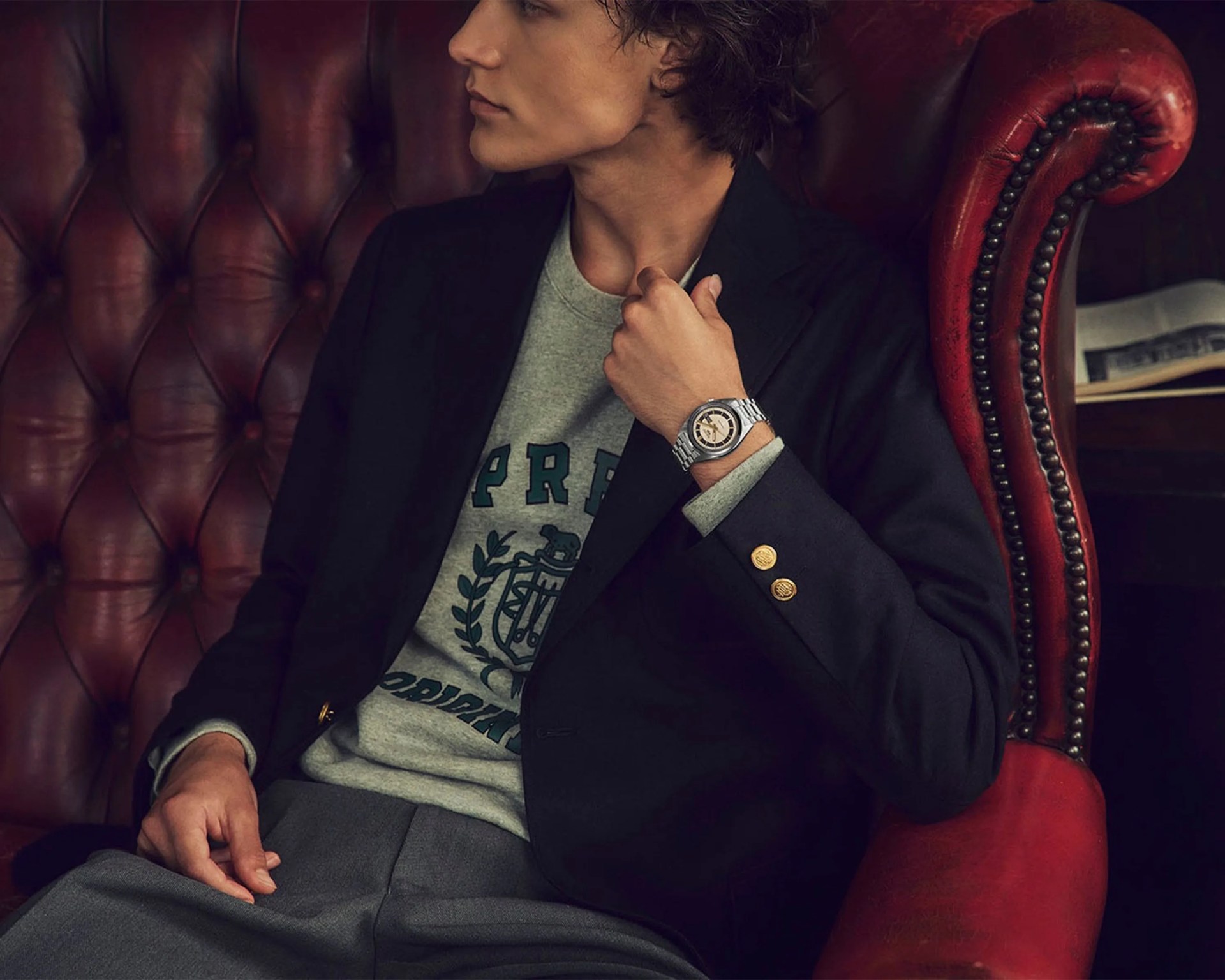 Person wearing a silver wristwatch with a round face, black blazer with gold buttons, and gray sweatshirt sitting on a red leather chair.