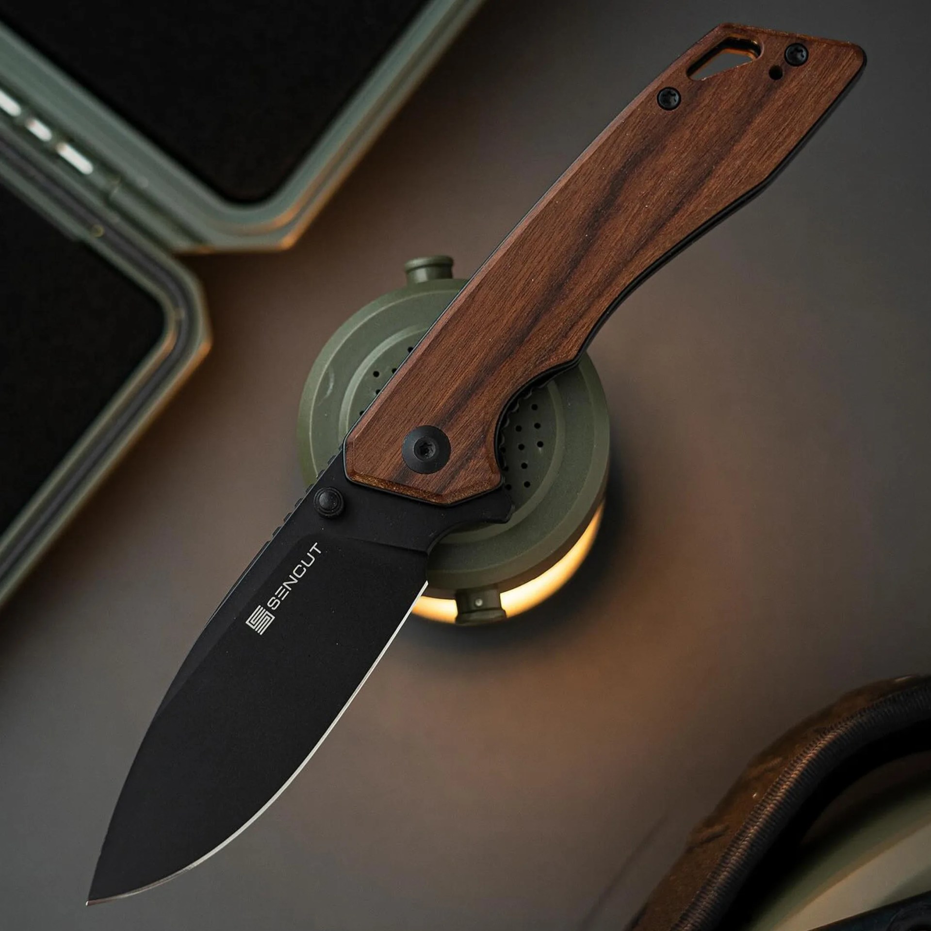Folding knife with black blade and wooden handle resting on a green lantern.