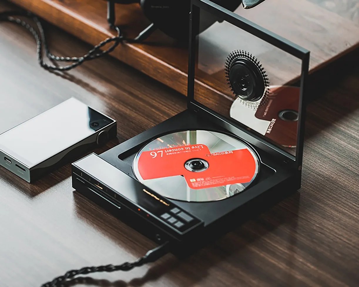 Black portable CD player with an open lid and a CD inside, placed on a wooden surface next to a silver device.