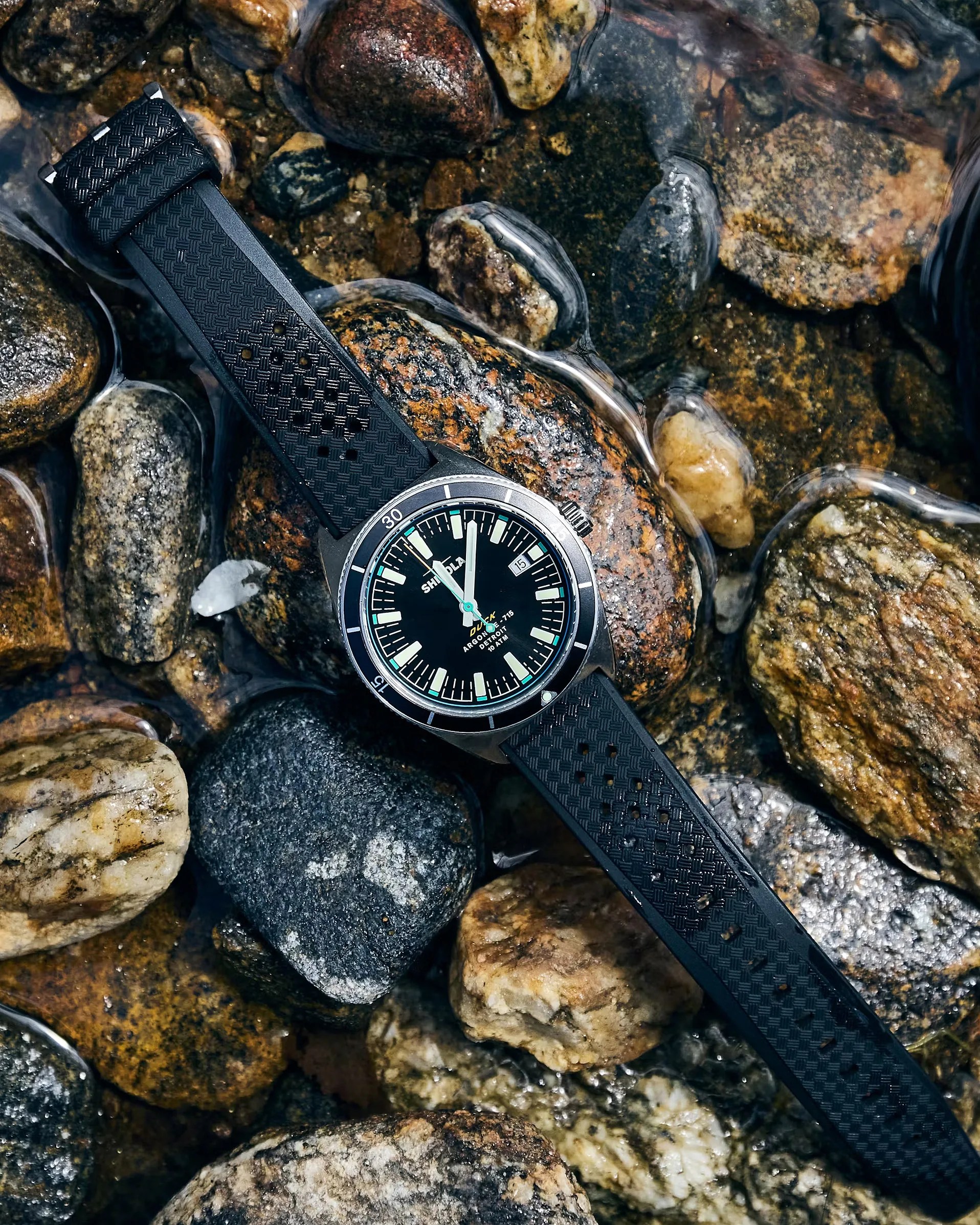 Black dive watch with luminous markers and textured black rubber strap resting on wet river rocks.