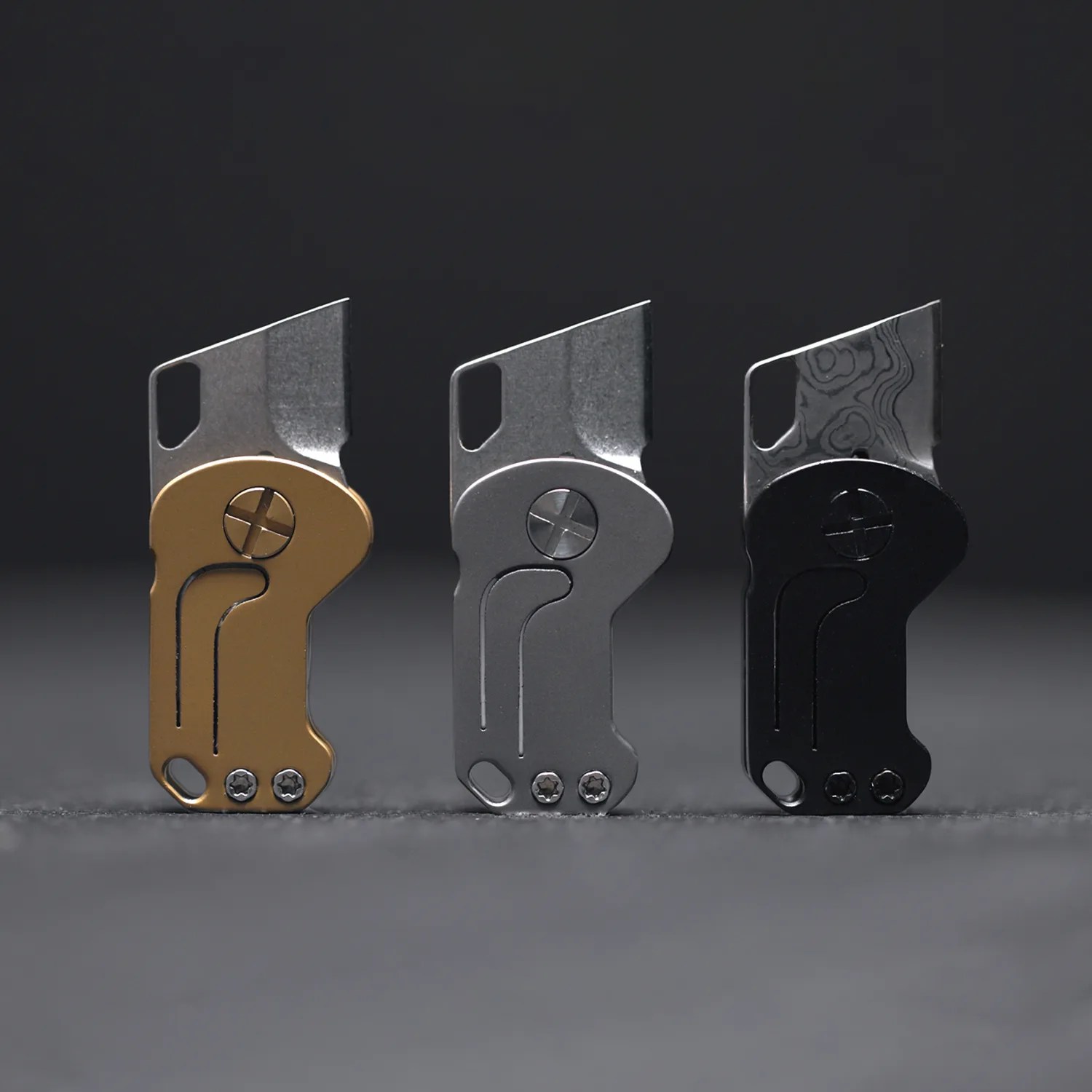 Three small folding utility knives in gold, silver, and black finishes standing upright on a gray surface.