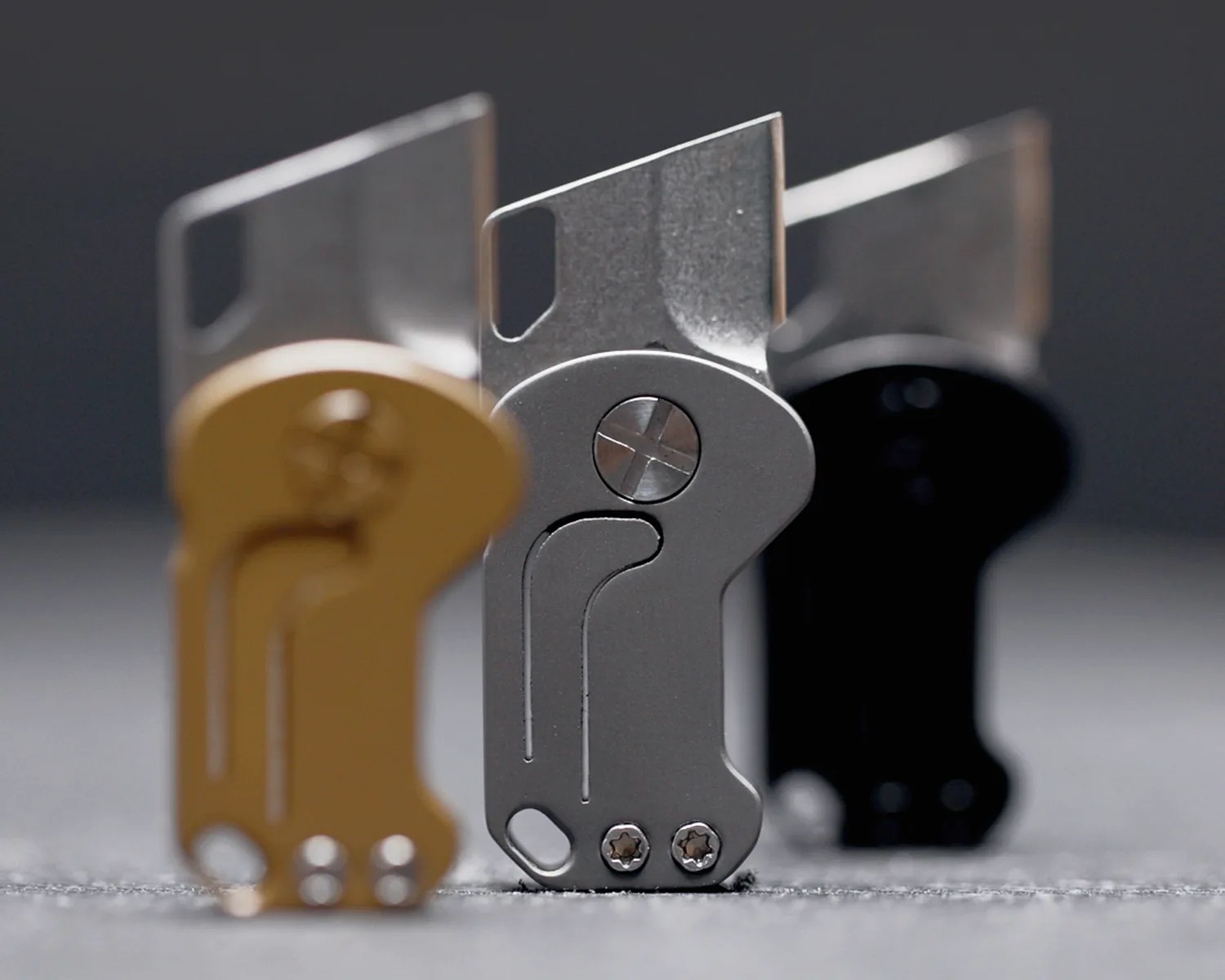 Three small folding utility knives in gold, silver, and black, standing upright on a gray surface.