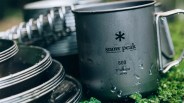 Stacked gray titanium camping mugs with "snow peak" logo and water droplets on green moss.