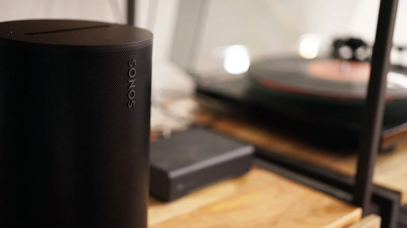 Black Sonos wireless speaker with mesh grille on a wooden surface near a turntable.
