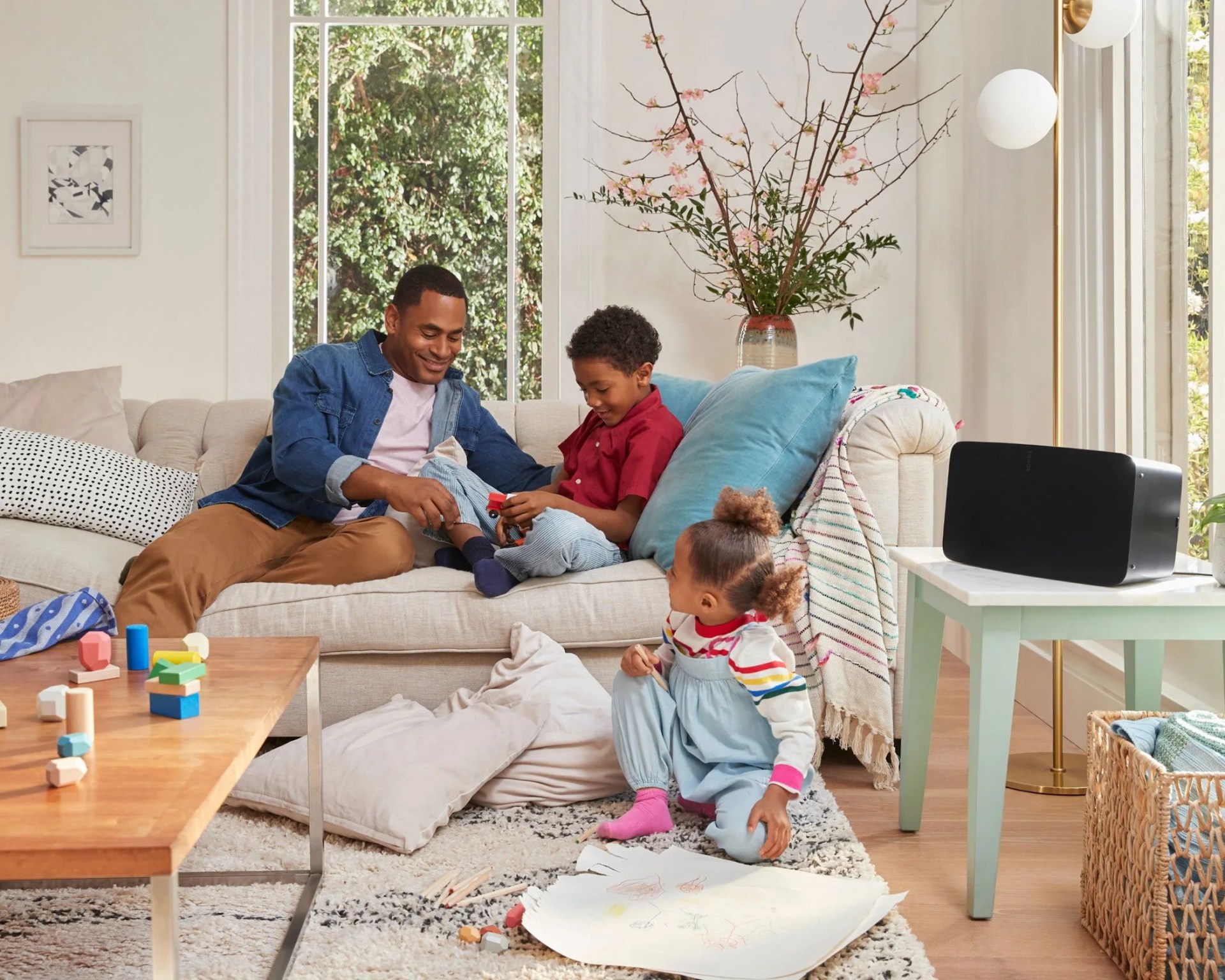 Living room with a beige sofa, a man and two children playing, a wooden coffee table with toys, and a black speaker on a side table.