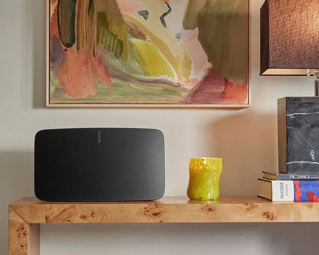 Black Sonos speaker on a wooden table next to a yellow glass and stacked books under an abstract painting.