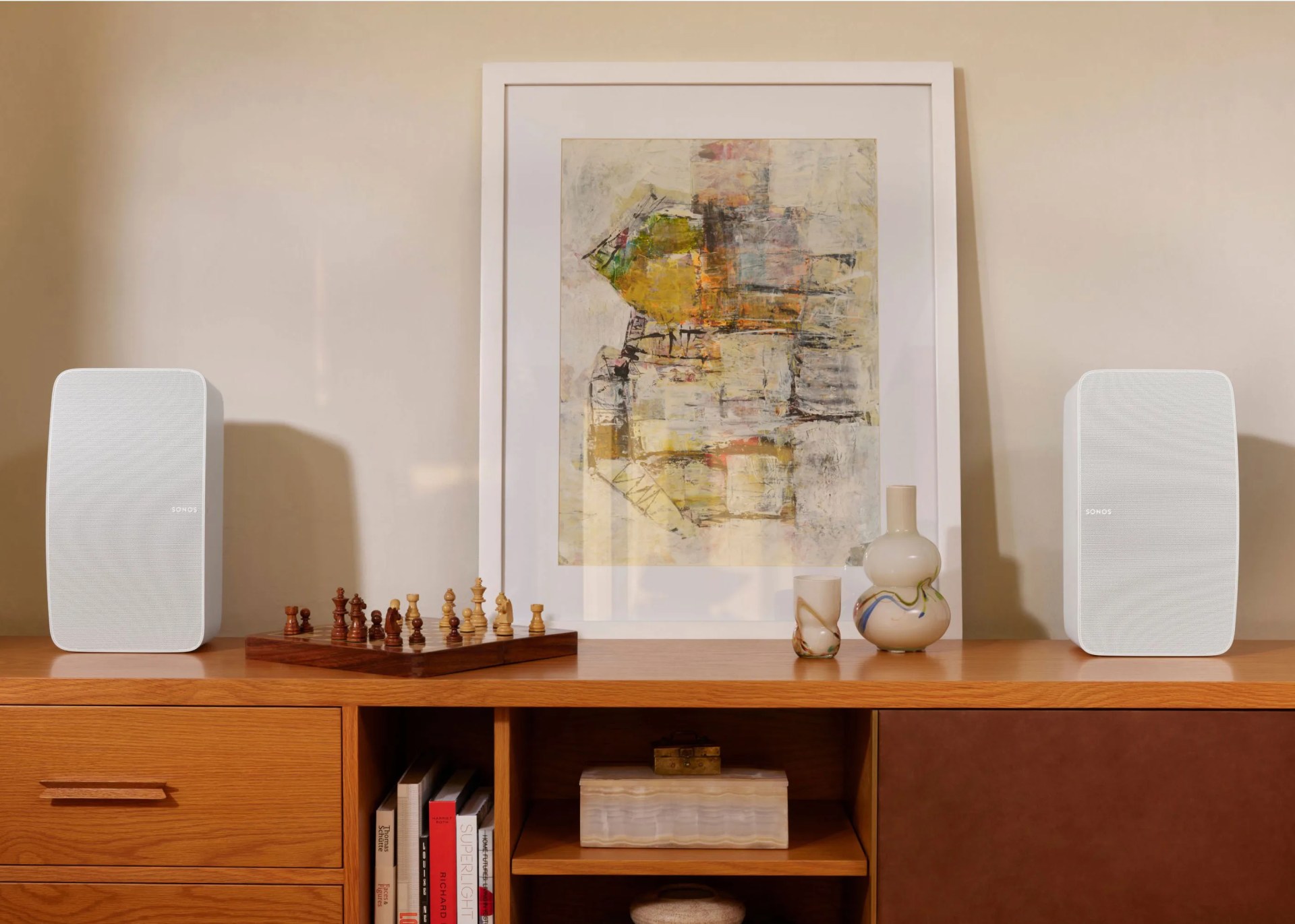 Pair of white Sonos speakers on a wooden cabinet with a chessboard, abstract framed art, and decorative vases.