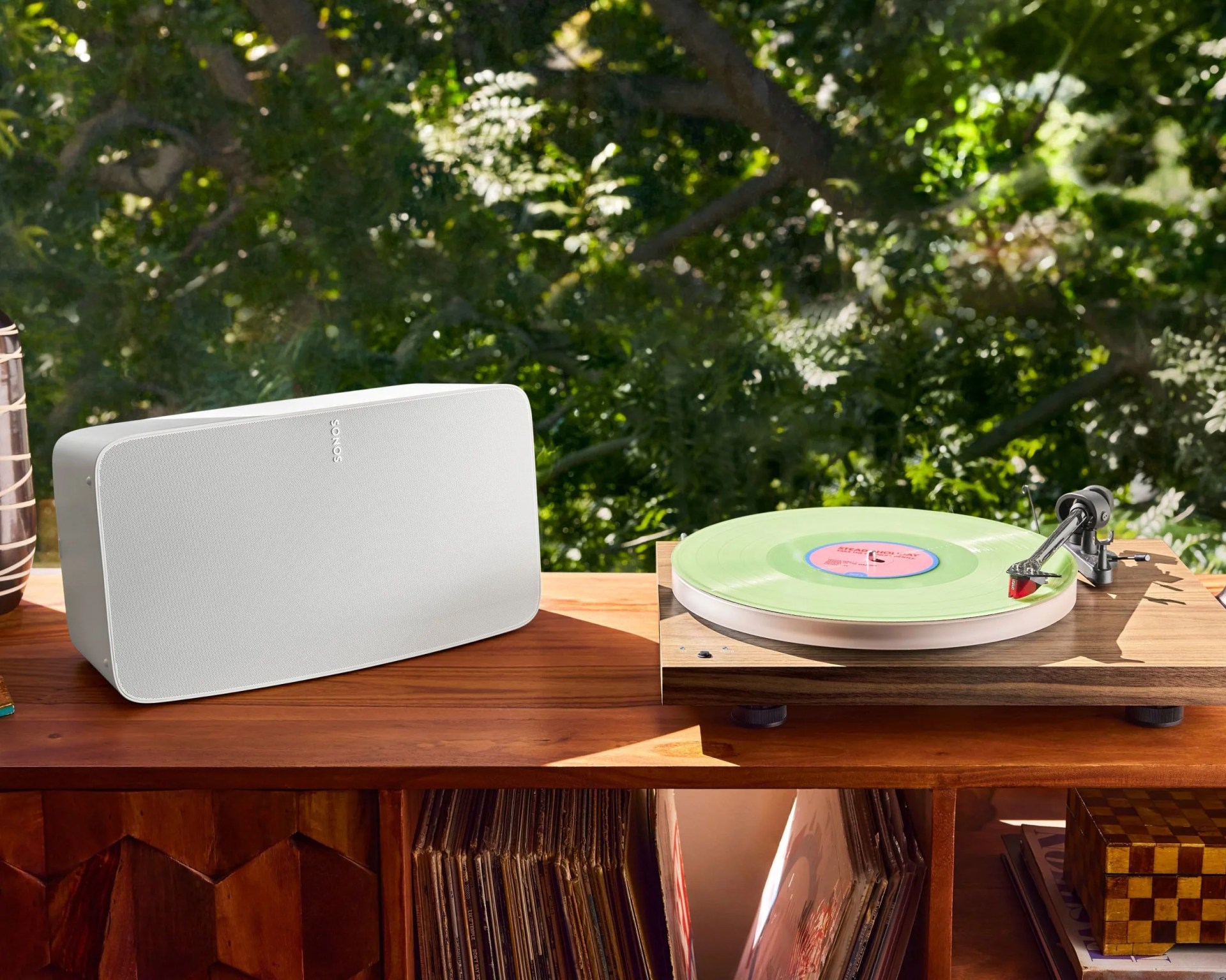 White Sonos speaker and wooden turntable with a green vinyl record on a wooden shelf with vinyl records below.