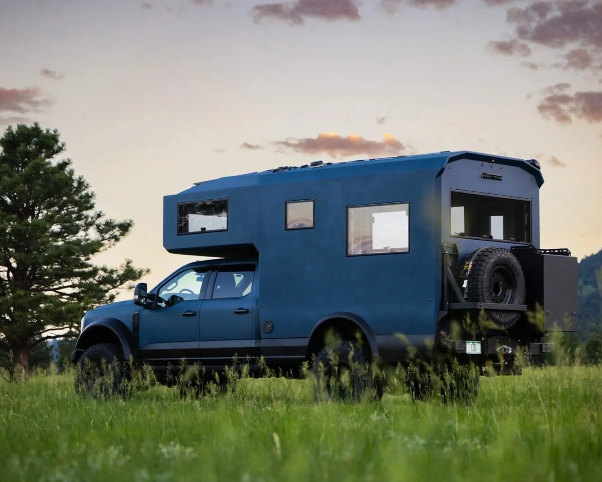 Blue off-road camper truck with large windows and spare tire mounted on the back in a grassy field.