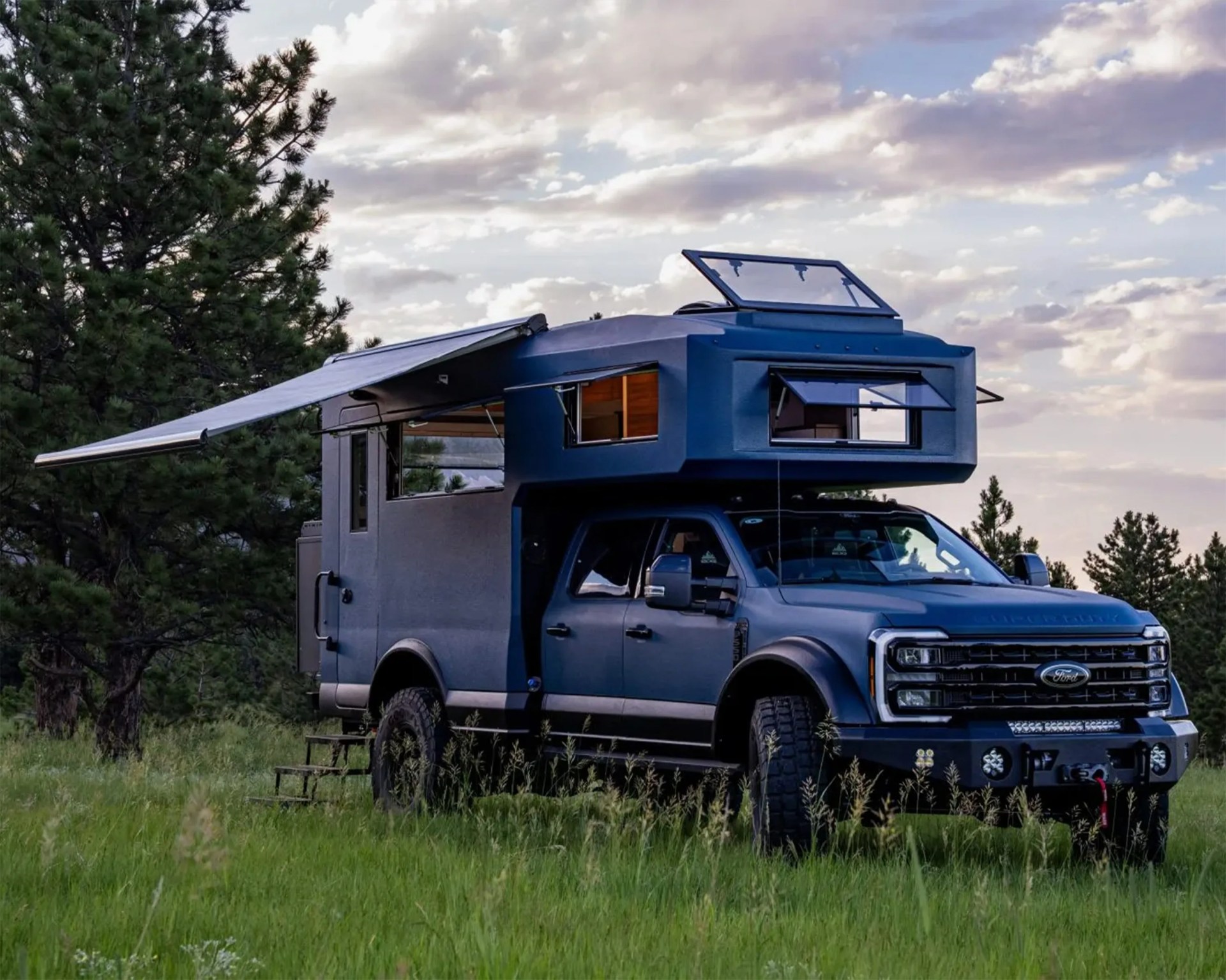Blue off-road camper truck with extended awning and open windows parked on grassy field near trees.