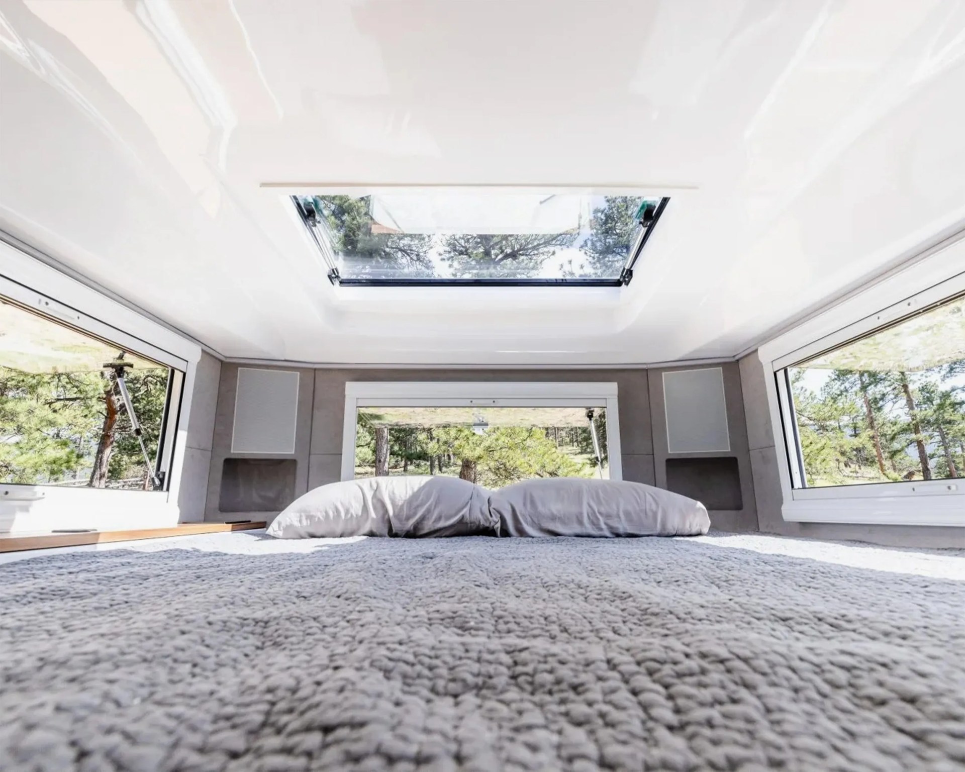 Bed with gray textured blanket and two pillows under a skylight and three large windows showing trees outside.