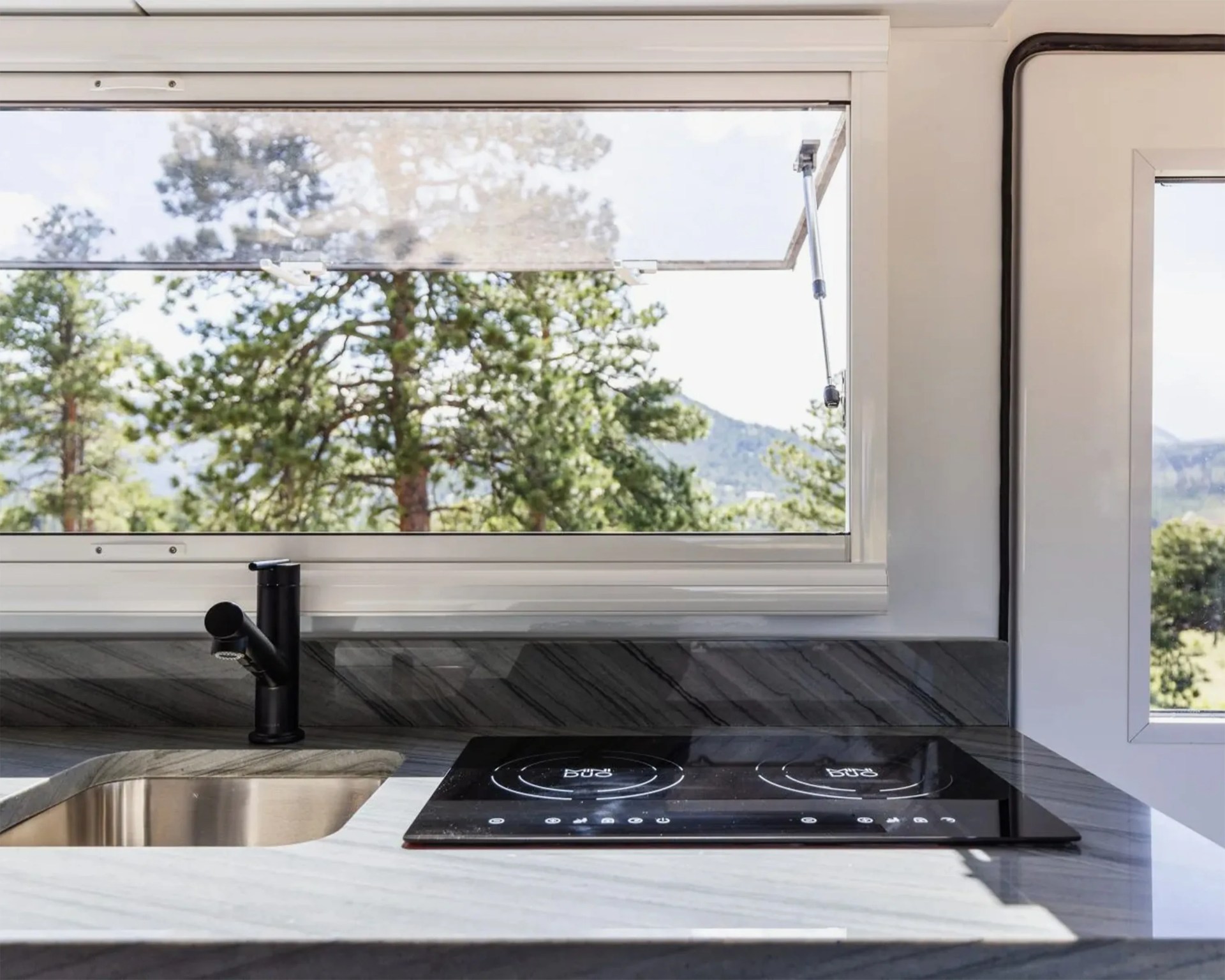 Marble countertop kitchen sink with black faucet and black electric cooktop under a window showing trees outside.