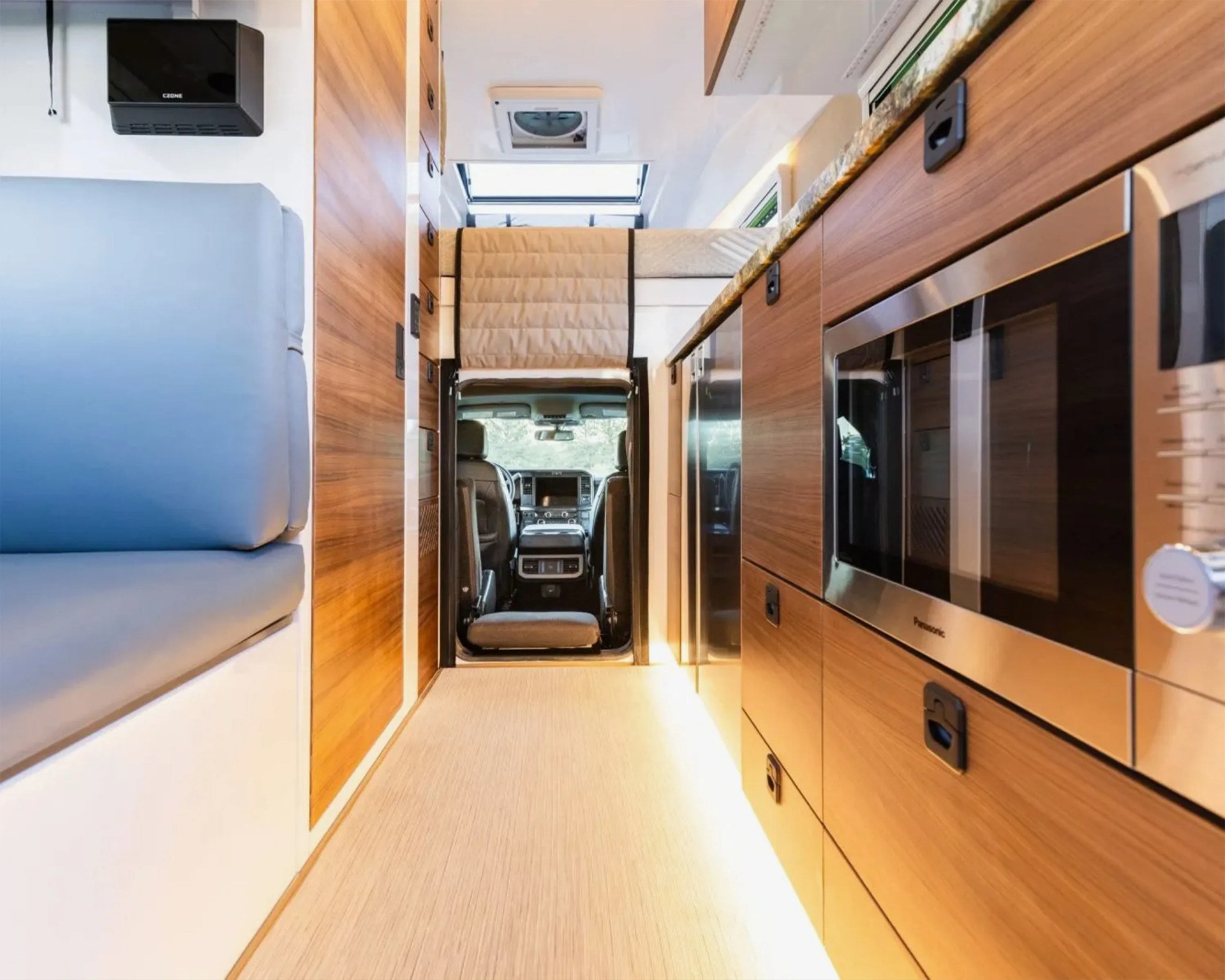 Interior aisle view of a camper van with wood cabinetry, a built-in microwave, and a blue cushioned bench.
