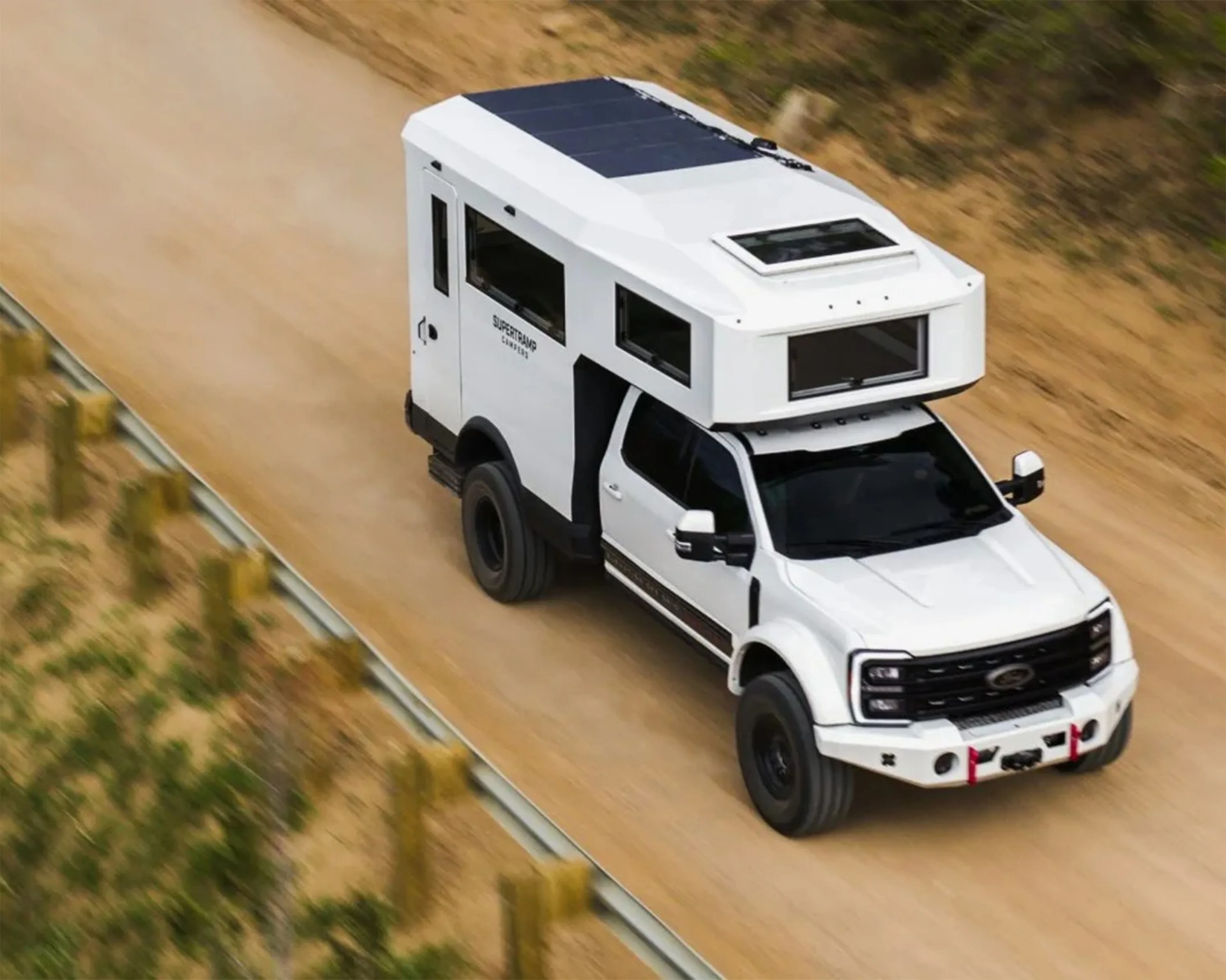 White off-road camper truck with black trim and solar panels driving on a dirt road.