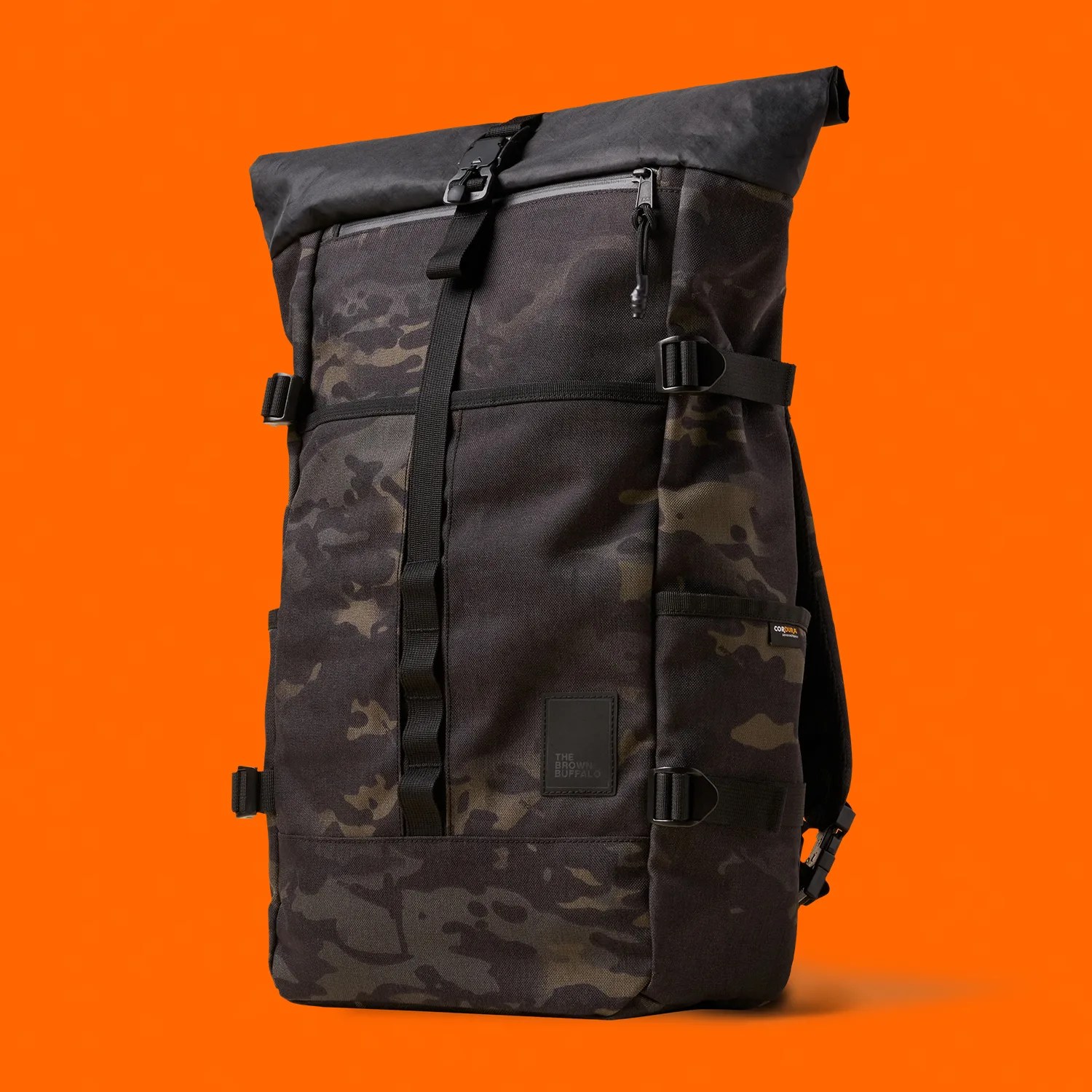 Black and dark camo roll-top backpack with front buckle and side compression straps against an orange background.