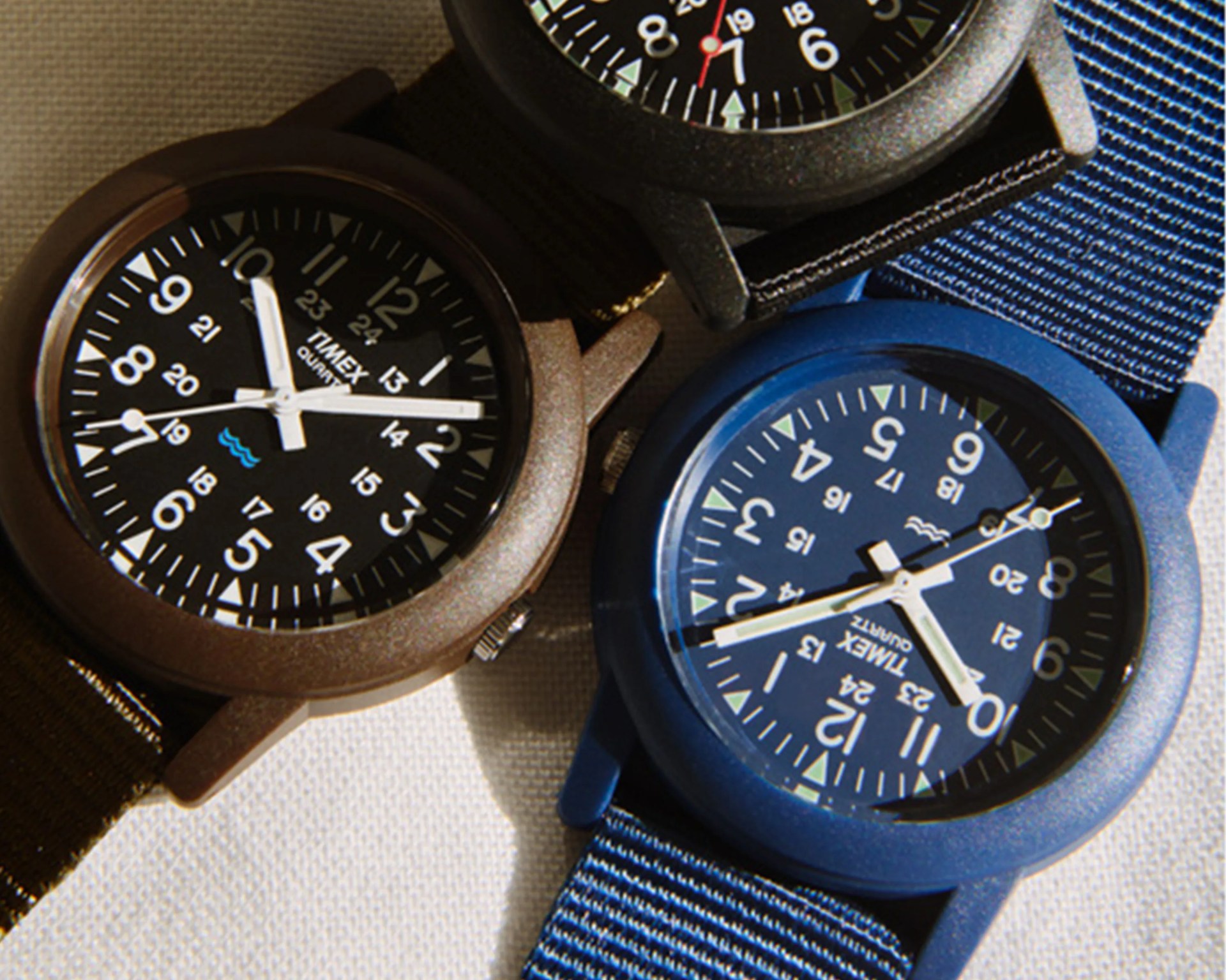 Three Timex wristwatches with black, brown, and blue cases and nylon straps on a light fabric surface.