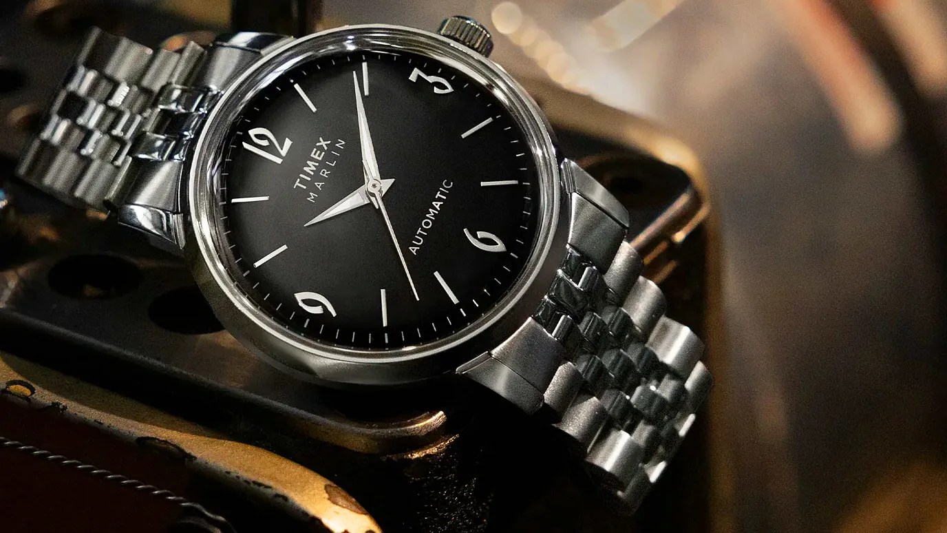 Silver Timex Marlin automatic wristwatch with black dial and metal link bracelet on a dark surface.