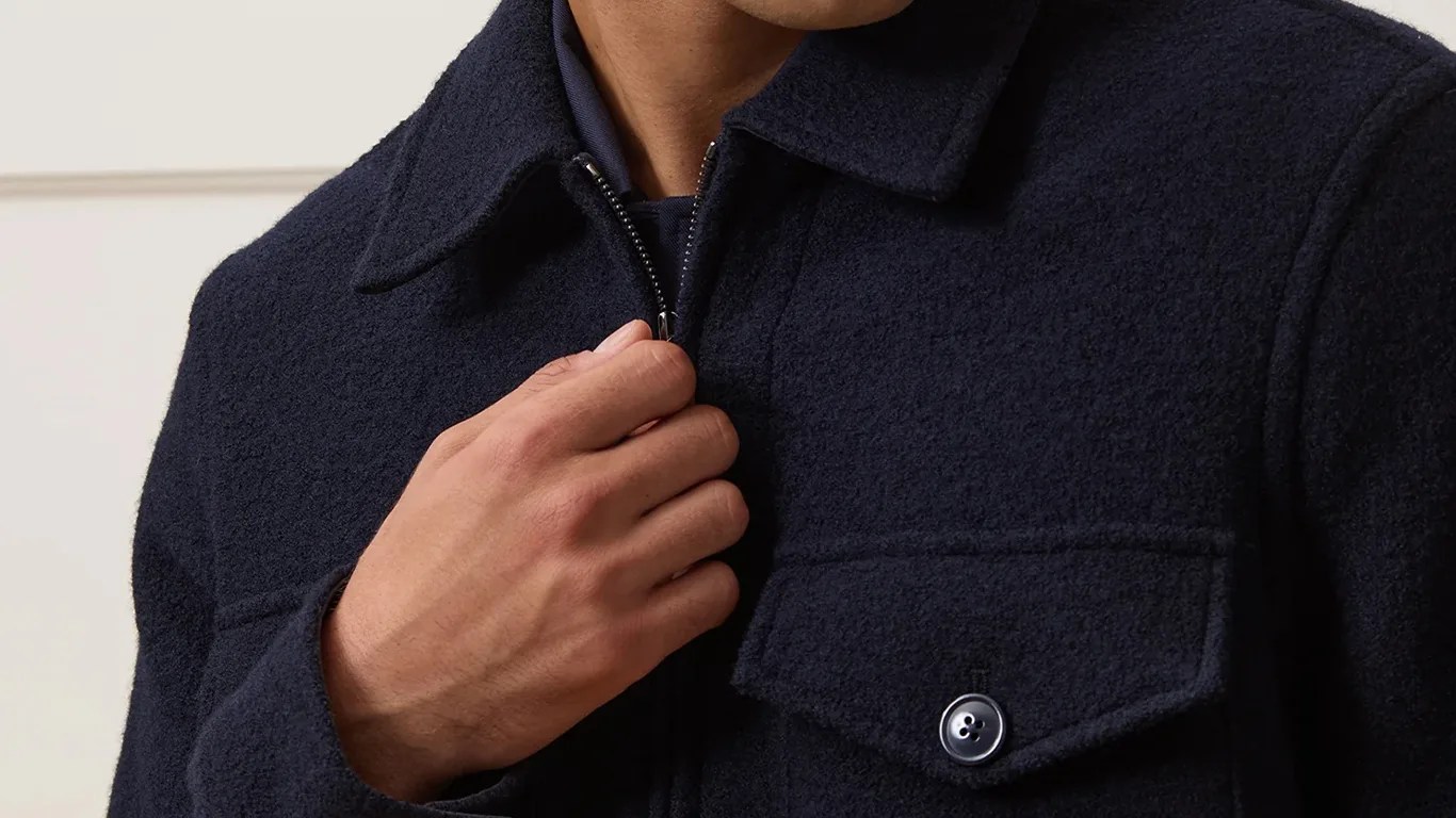 Close-up of a hand zipping up a Todd Snyder textured dark navy wool jacket with a buttoned chest pocket.