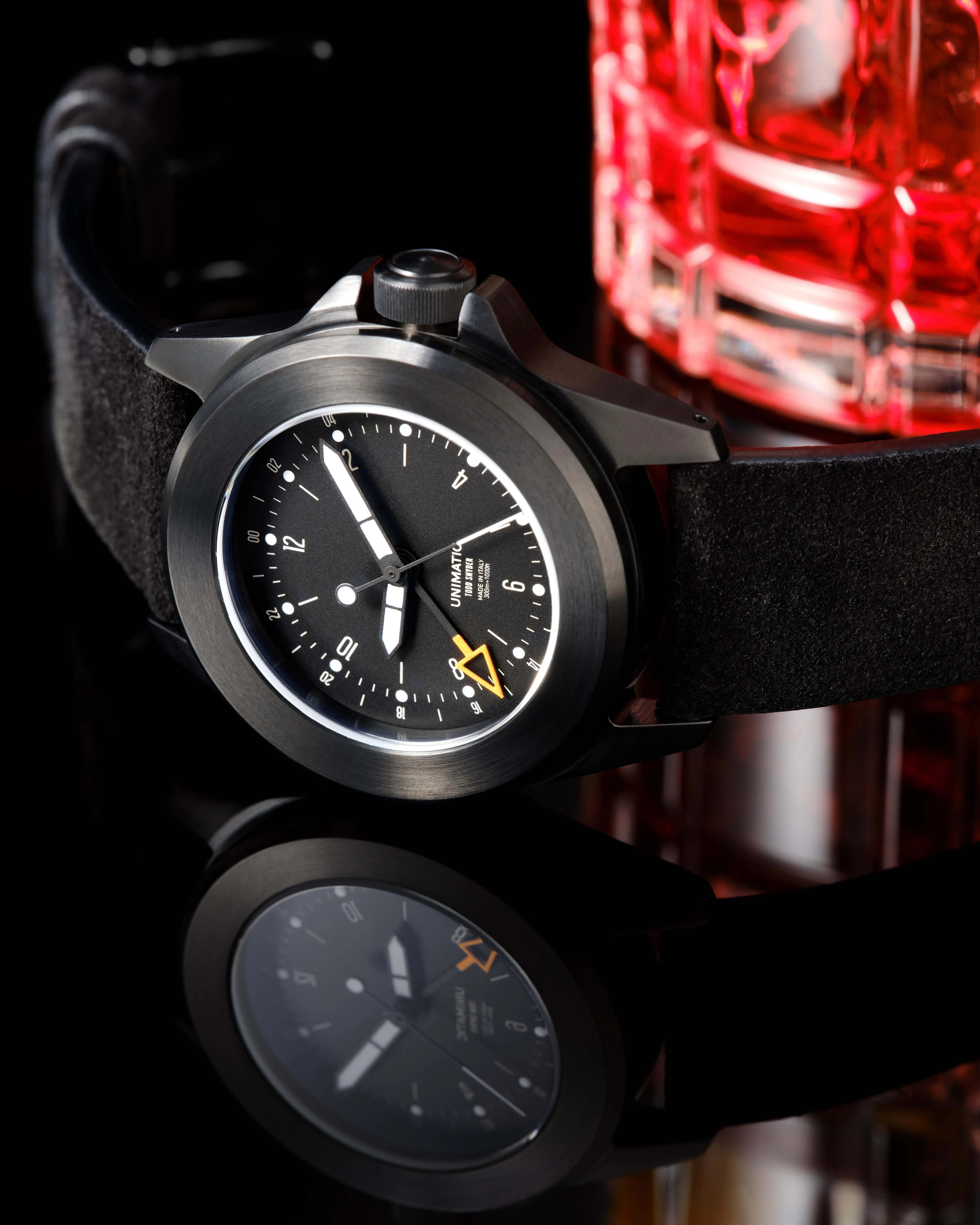 Black Unimatic wristwatch with brushed metal case, bold white hands, orange arrow-tipped hand, and black suede strap.