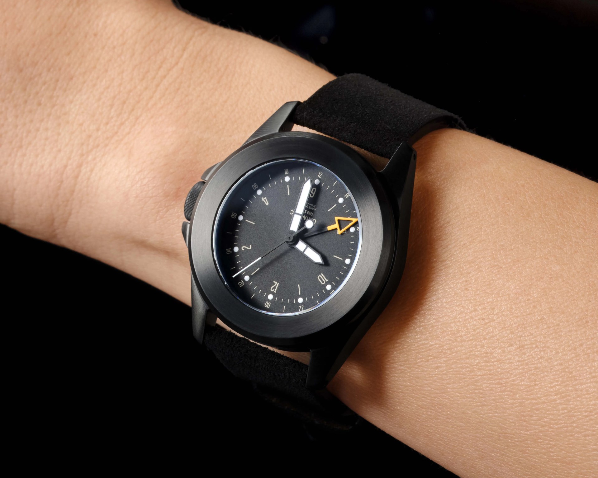 Black wristwatch with a matte black case, black dial, white hour and minute hands, and an orange arrow-tipped second hand on a black fabric strap.