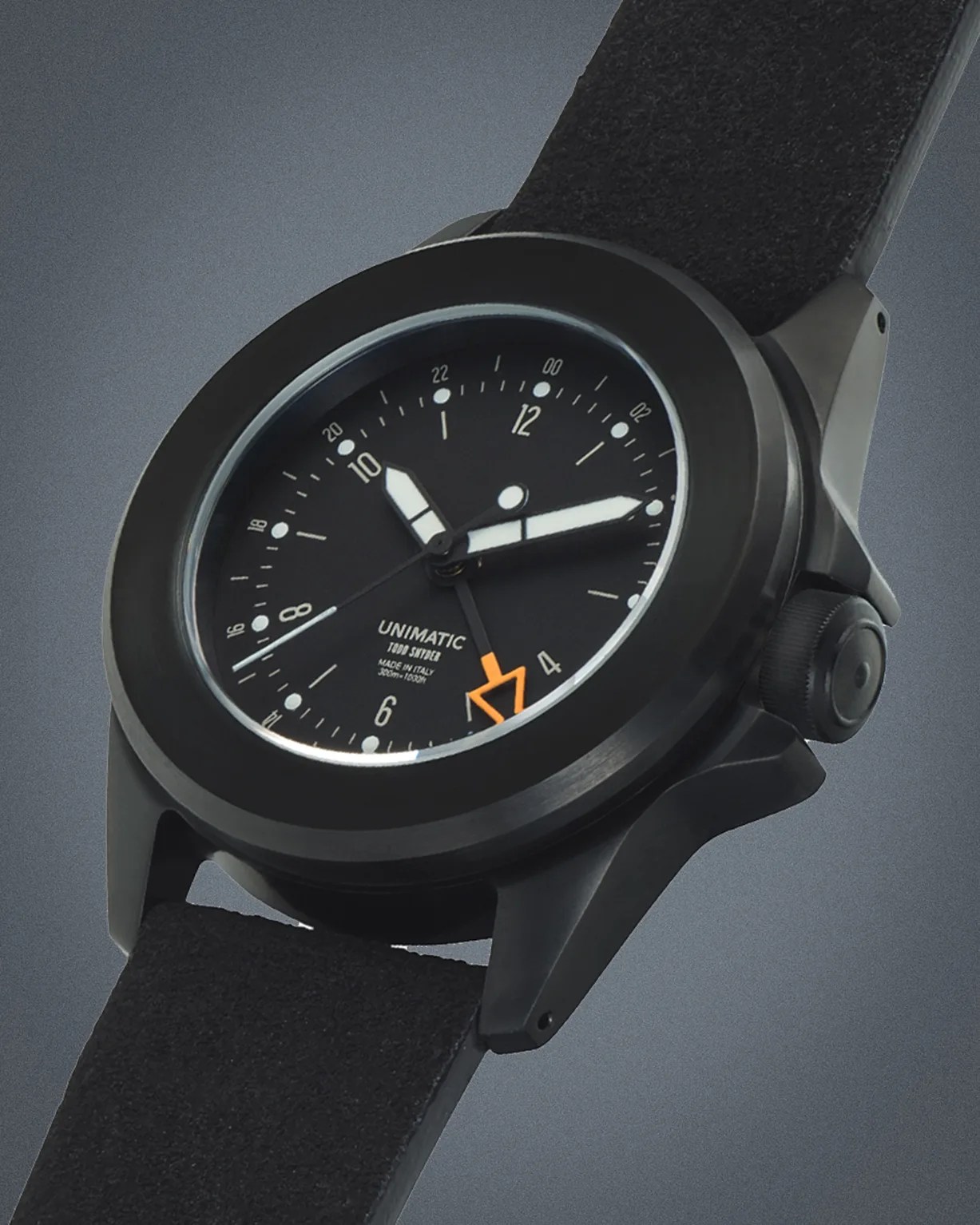 Black Unimatic wristwatch with matte finish, black dial, white hour markers, orange arrow second hand, and black suede strap.