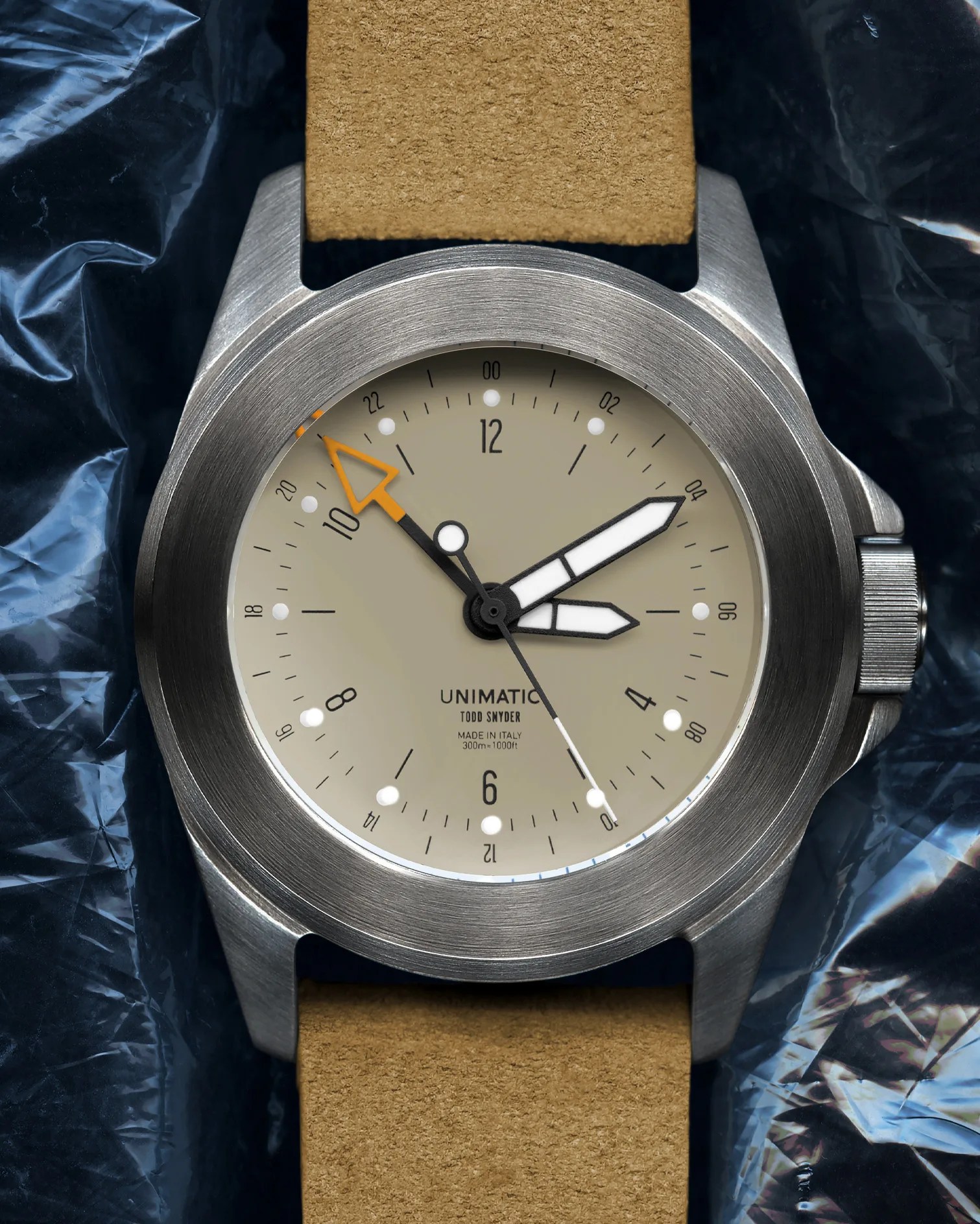 Unimatic wristwatch with beige dial, orange 24-hour hand, brushed stainless steel case, and tan suede strap.