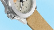 Close-up of a silver metal watch case with a beige suede strap on a light blue background.