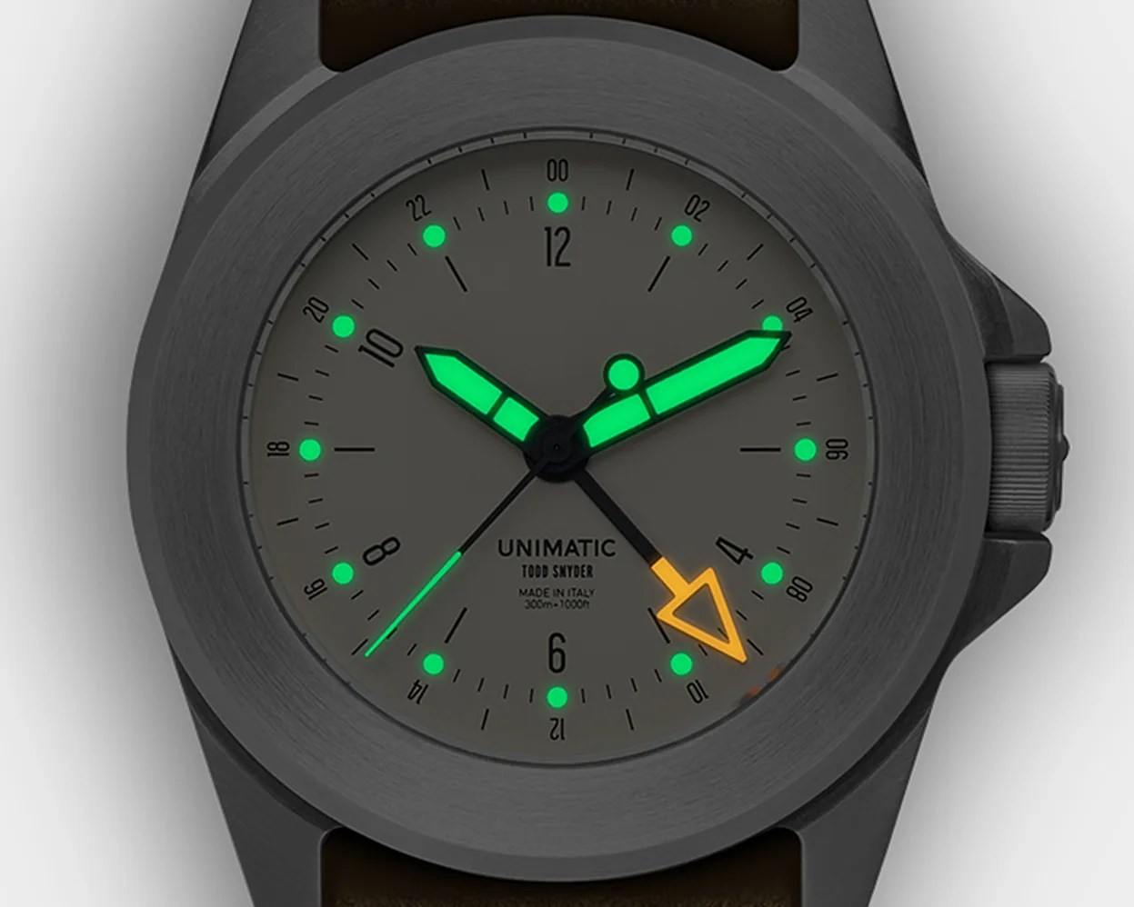 Close-up of a Unimatic Todd Snyder watch face with green and orange luminescent hands and markers on a gray dial.