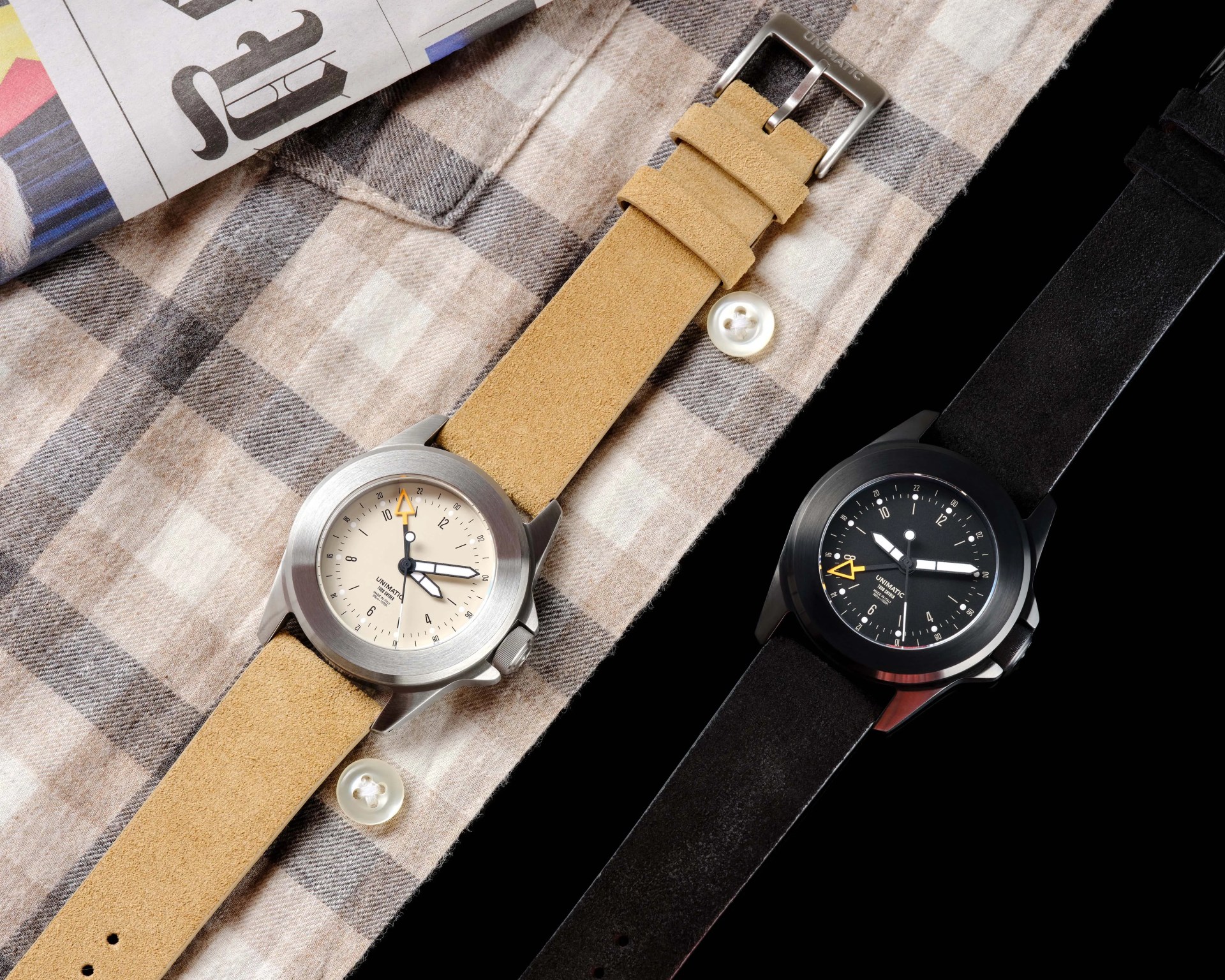 Two wristwatches with suede straps, one beige with a cream dial and one black with a black dial, placed on a plaid shirt and black surface.