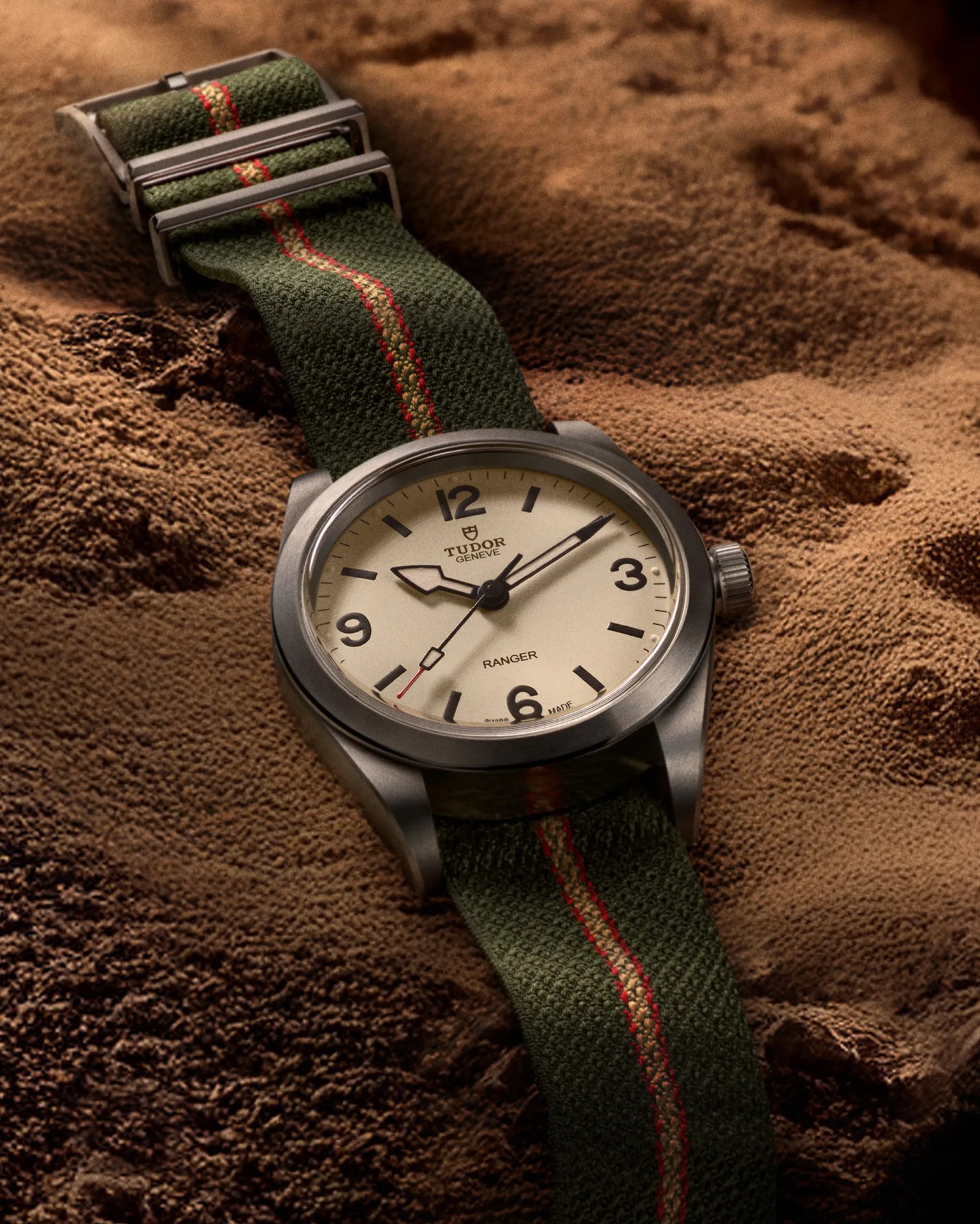 Tudor Ranger watch with beige dial and green fabric strap with red stripe on textured brown surface.