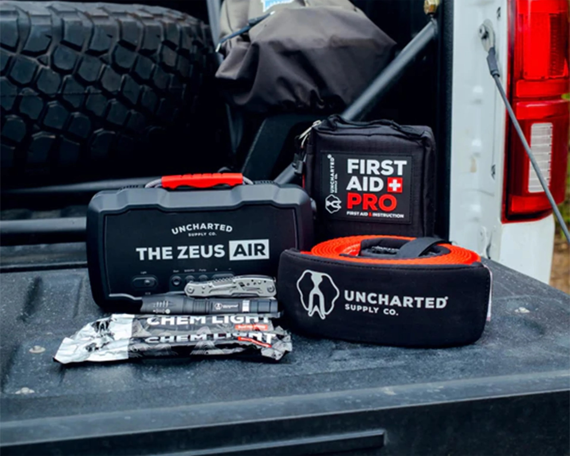 Emergency roadside kit including a black and red "The Zeus Air" inflator, first aid kit, tow strap, flashlight, multitool, and chem lights on a truck bed.