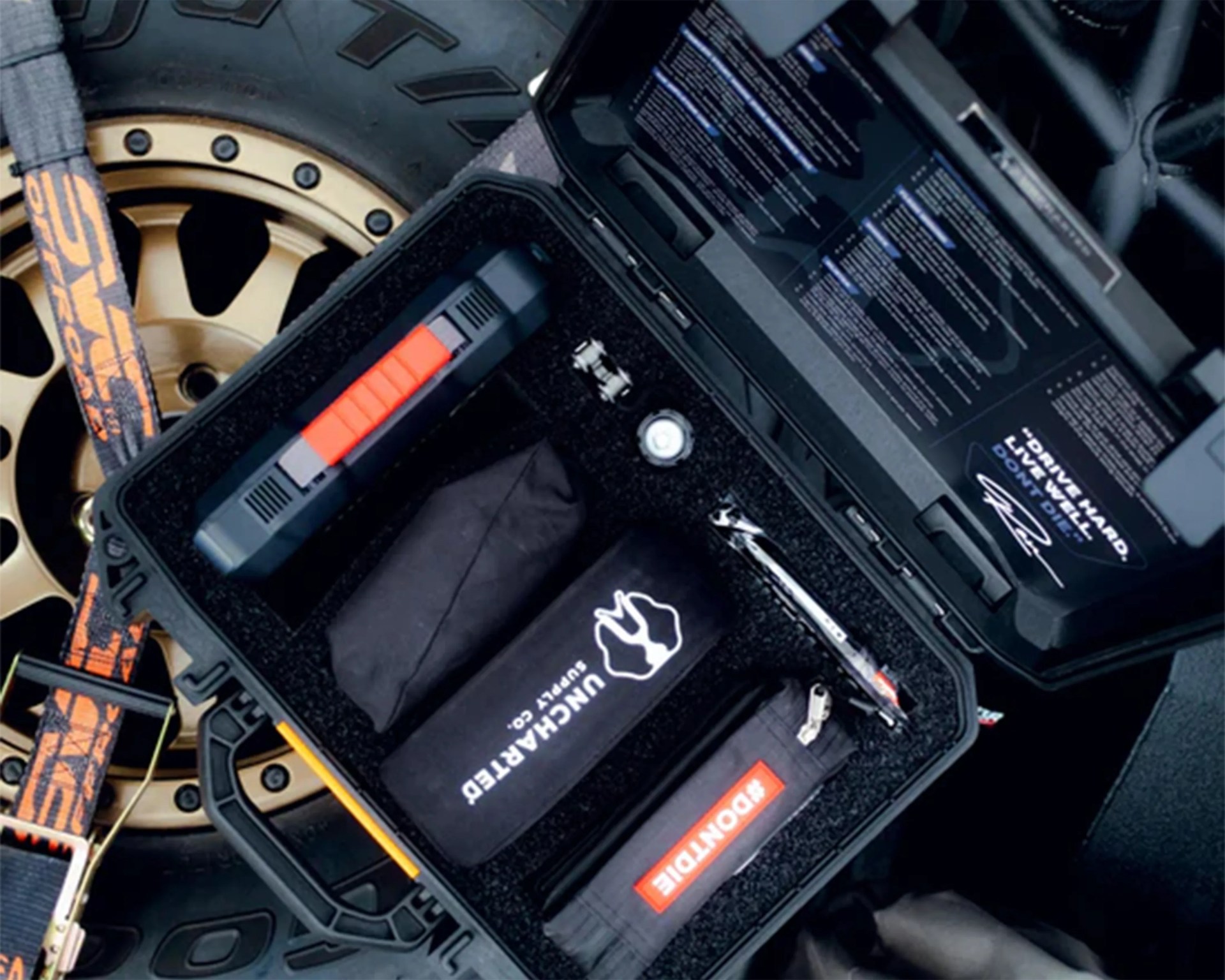 Black hard case with foam insert containing black pouches, a tool, and a device with a red grip, placed near a gold vehicle wheel.
