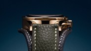 Close-up of a gold watch case with a green textured leather strap and white stitching against a dark blue background.
