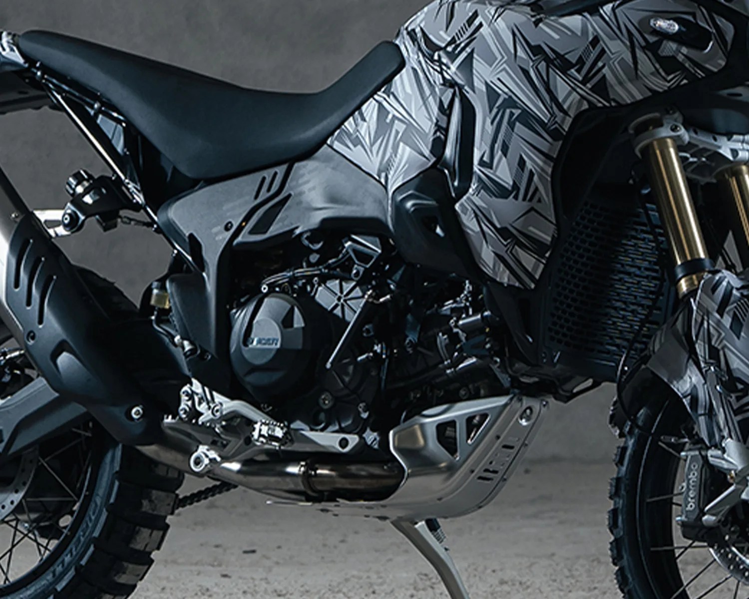 Close-up of a motorcycle engine and seat with a black and gray geometric patterned body and knobby tires.
