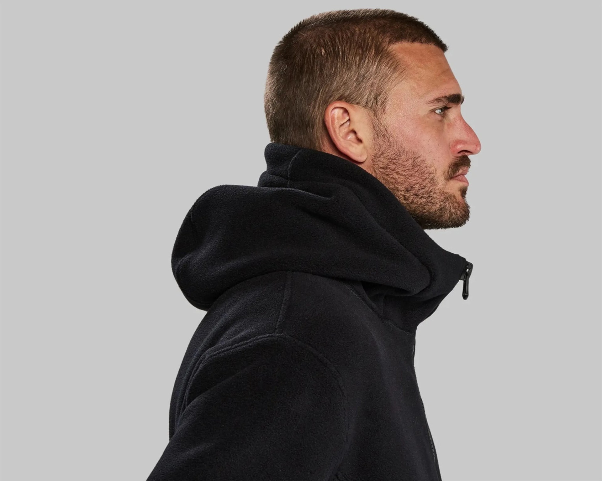 Side profile of a man wearing a black fleece jacket with a high collar and hood against a gray background.