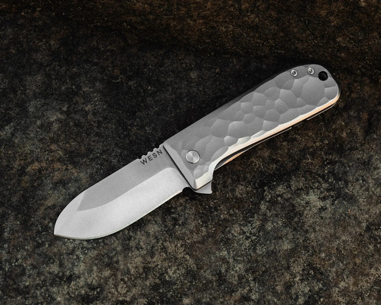 Folding knife with a textured silver handle and a wide matte blade engraved with "WESN" on a dark stone surface.