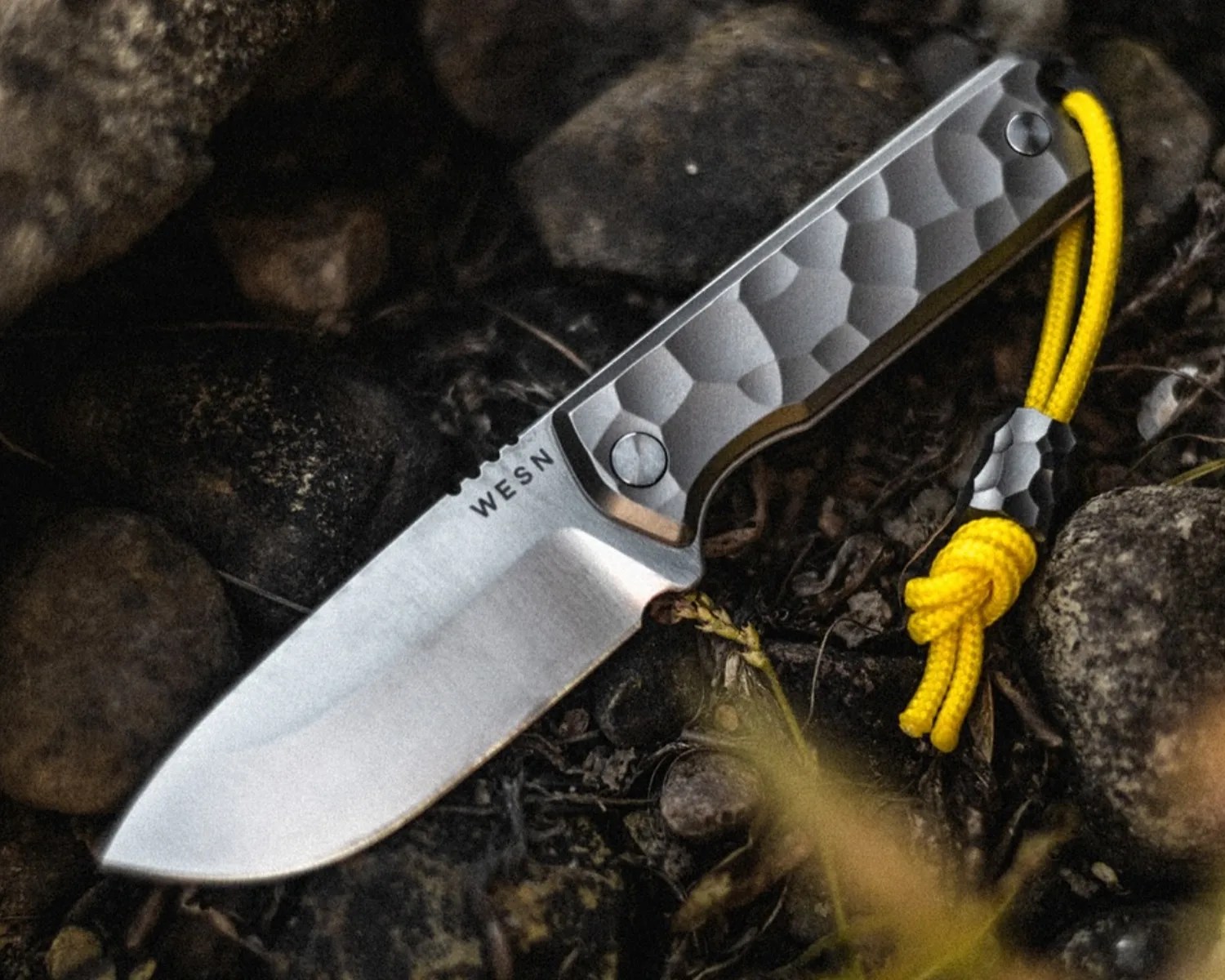 Fixed-blade knife with a textured metal handle and yellow lanyard, lying on dark rocks and soil.
