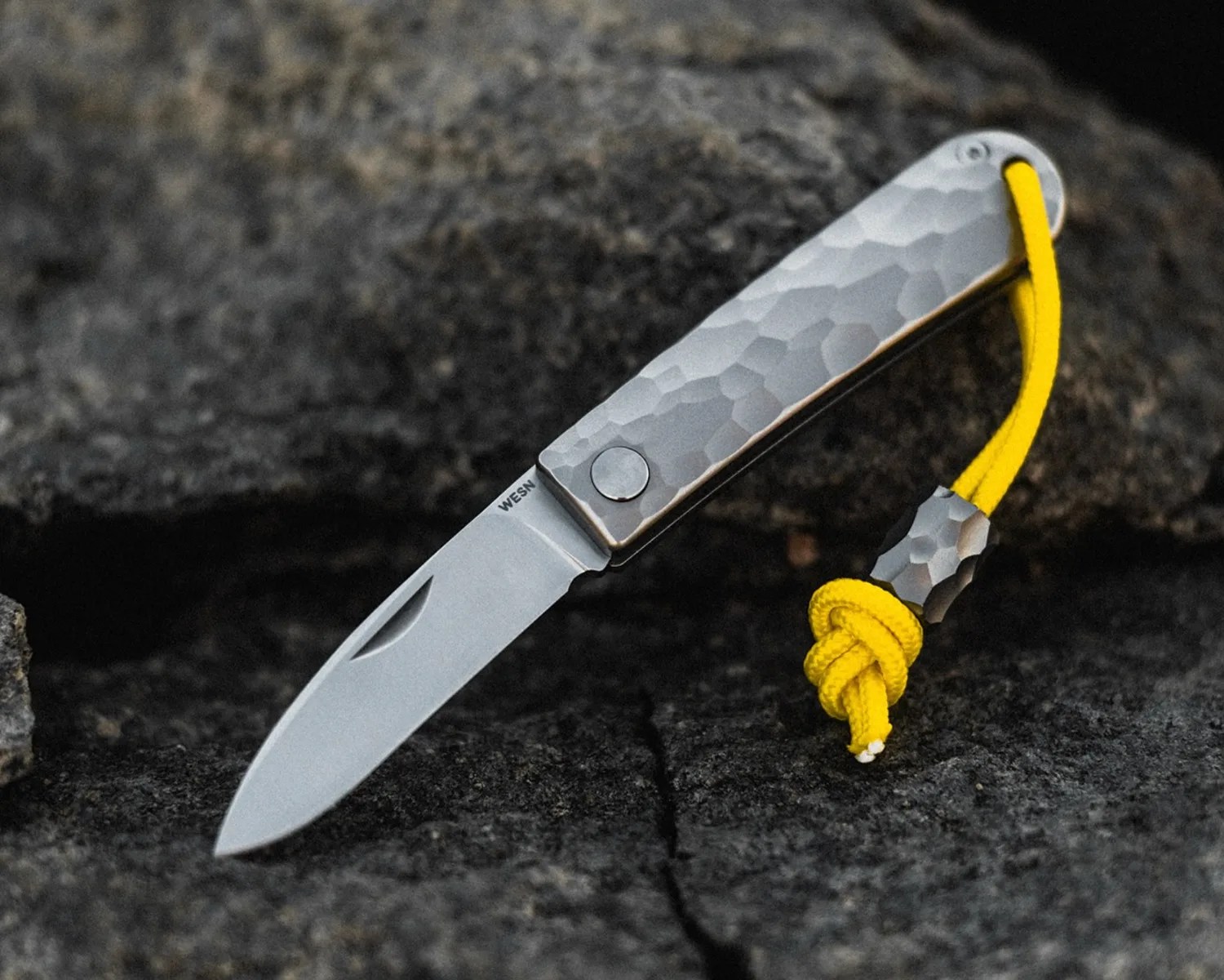 Folding knife with hammered metal handle and yellow lanyard on dark textured rock surface.