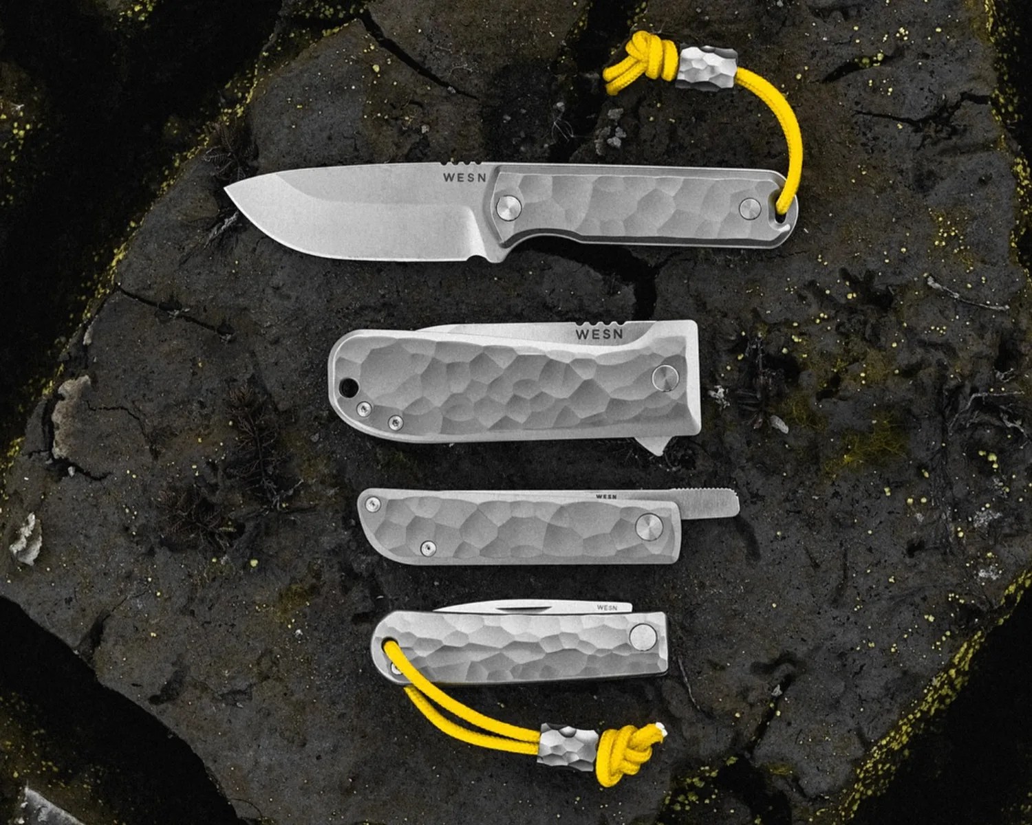 Four silver folding knives with textured handles and yellow lanyards, arranged on dark rock.