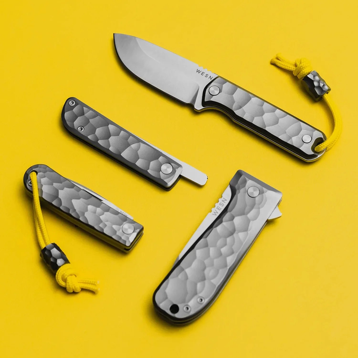 Four titanium folding knives with hammered texture and yellow lanyards on a yellow background.