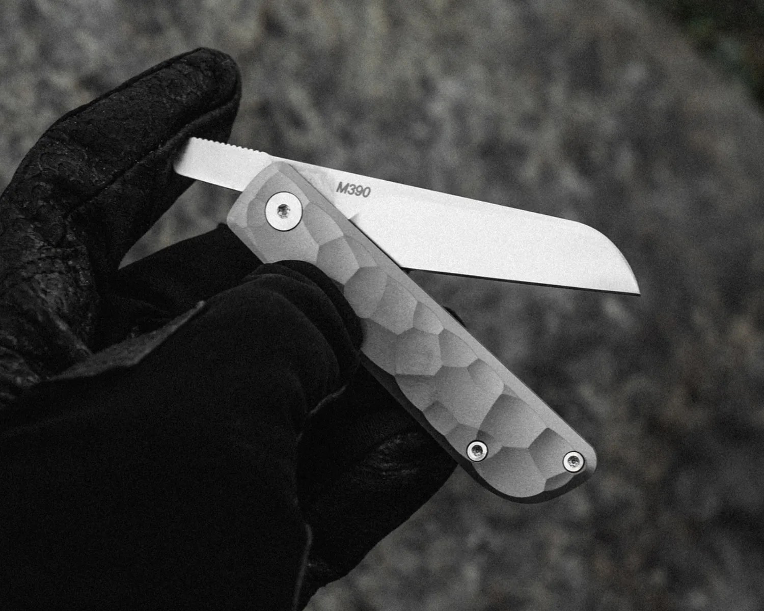 Hand in black glove holding a folding knife with a textured gray handle and a straight-edged blade marked "M390".