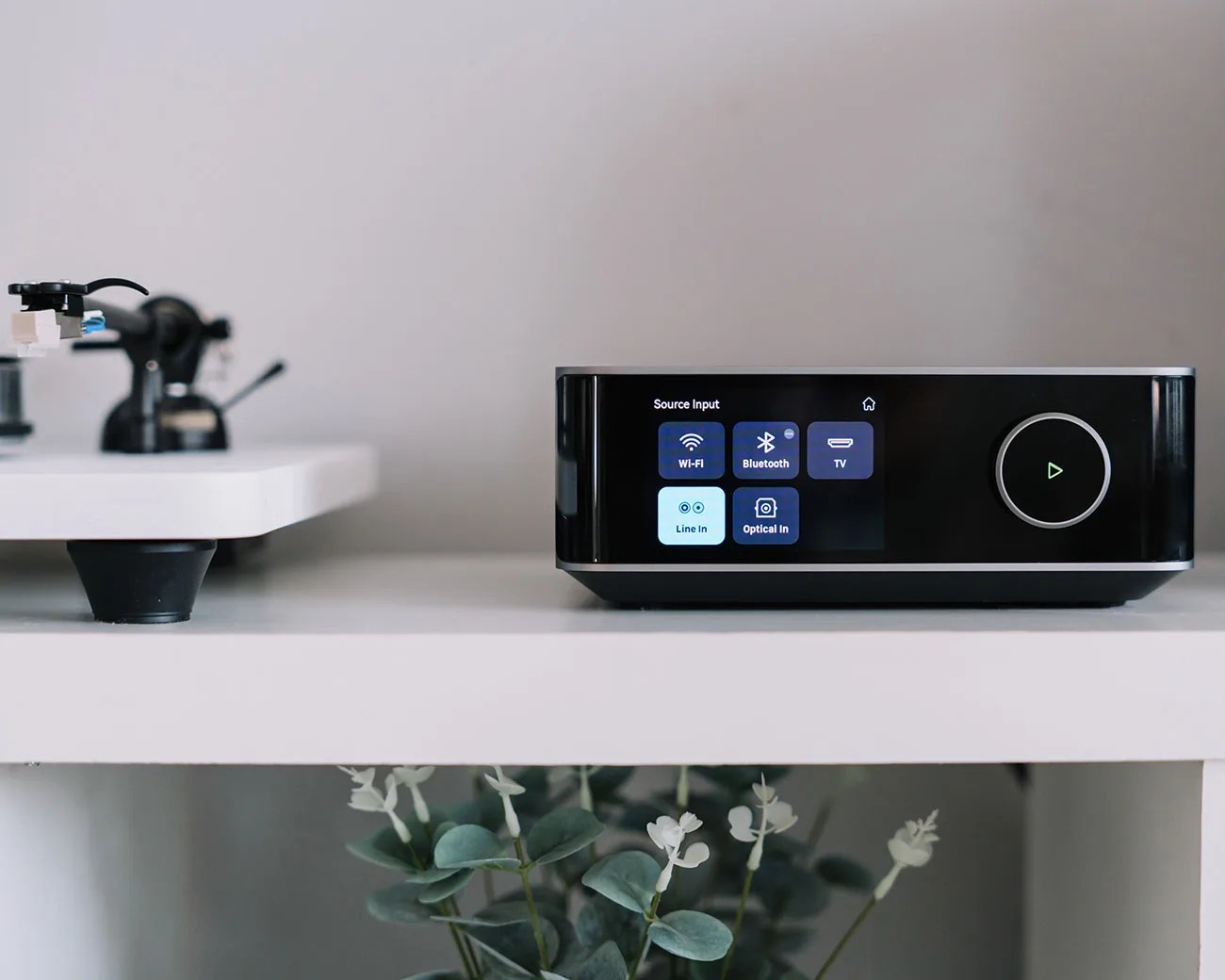 Black audio device with touchscreen showing source input options on a white shelf next to a turntable.