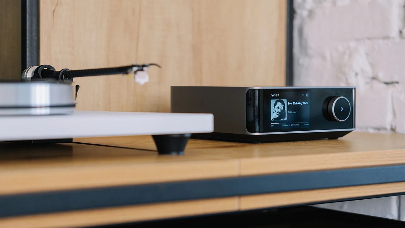 Black and silver digital music player with display screen and control knob on wooden shelf next to a white turntable.
